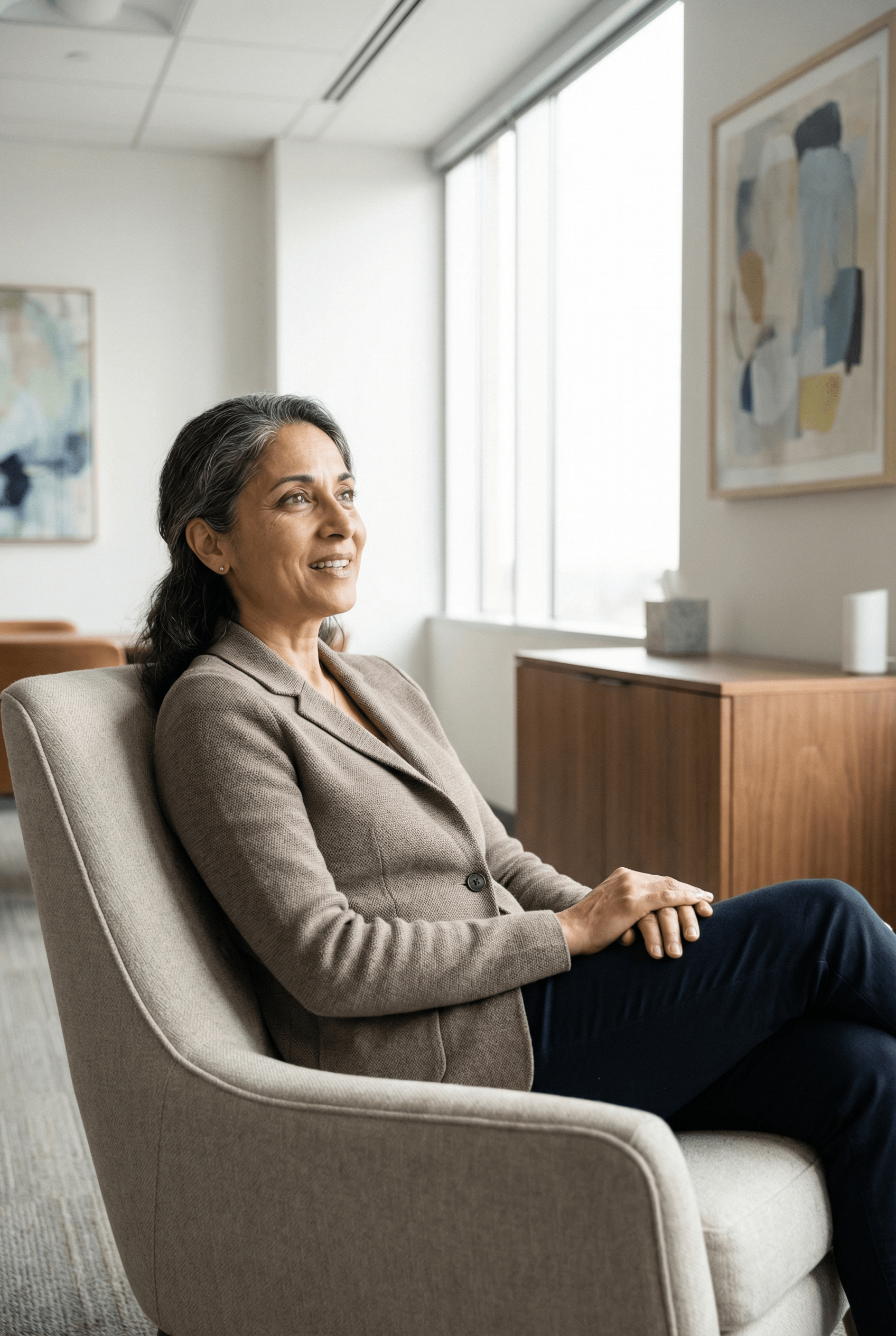 Woman in blazer seated in armchair, looking out a window in an office setting.