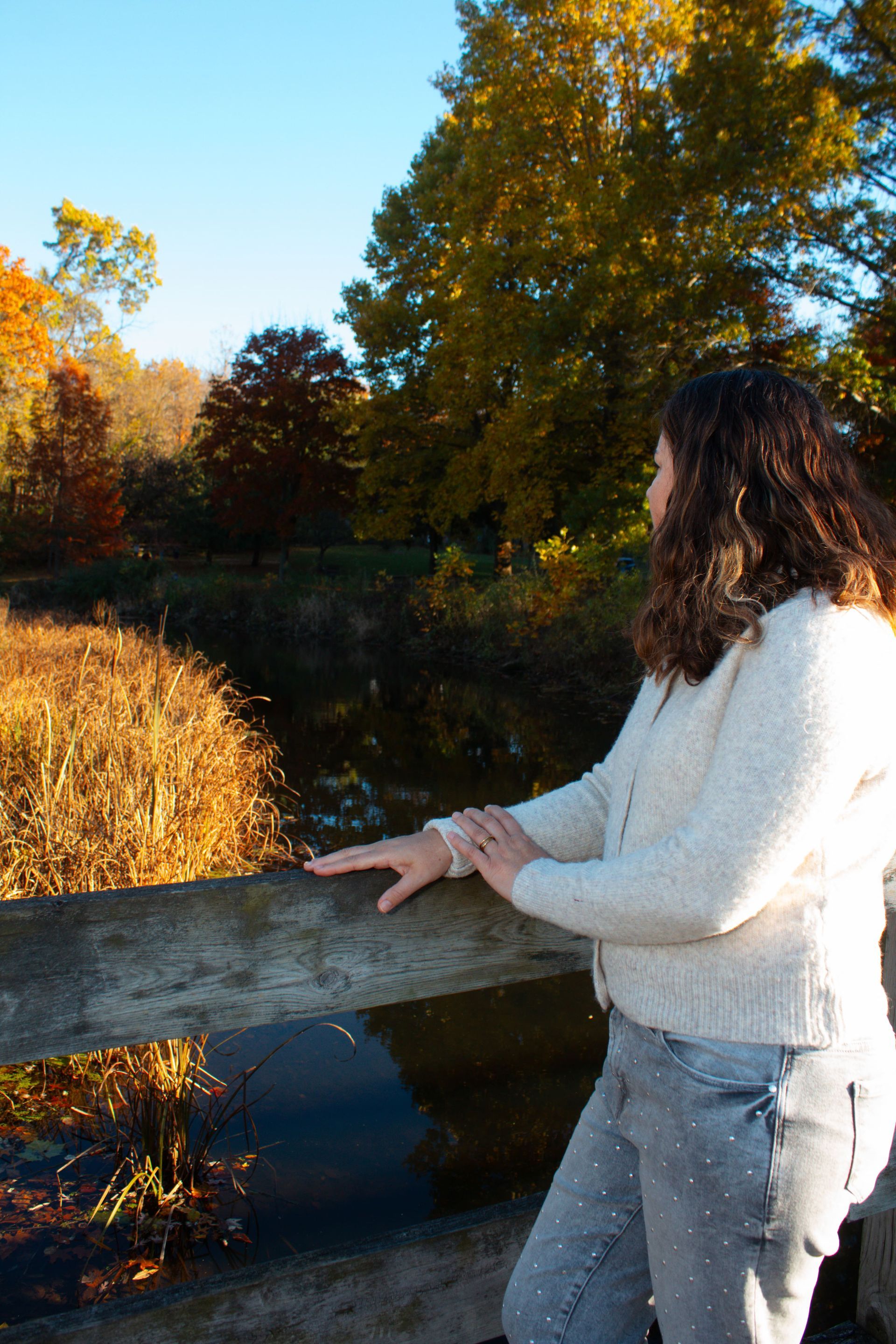 Woman in white sweater leans on a wooden railing, gazing at a pond surrounded by autumn trees.