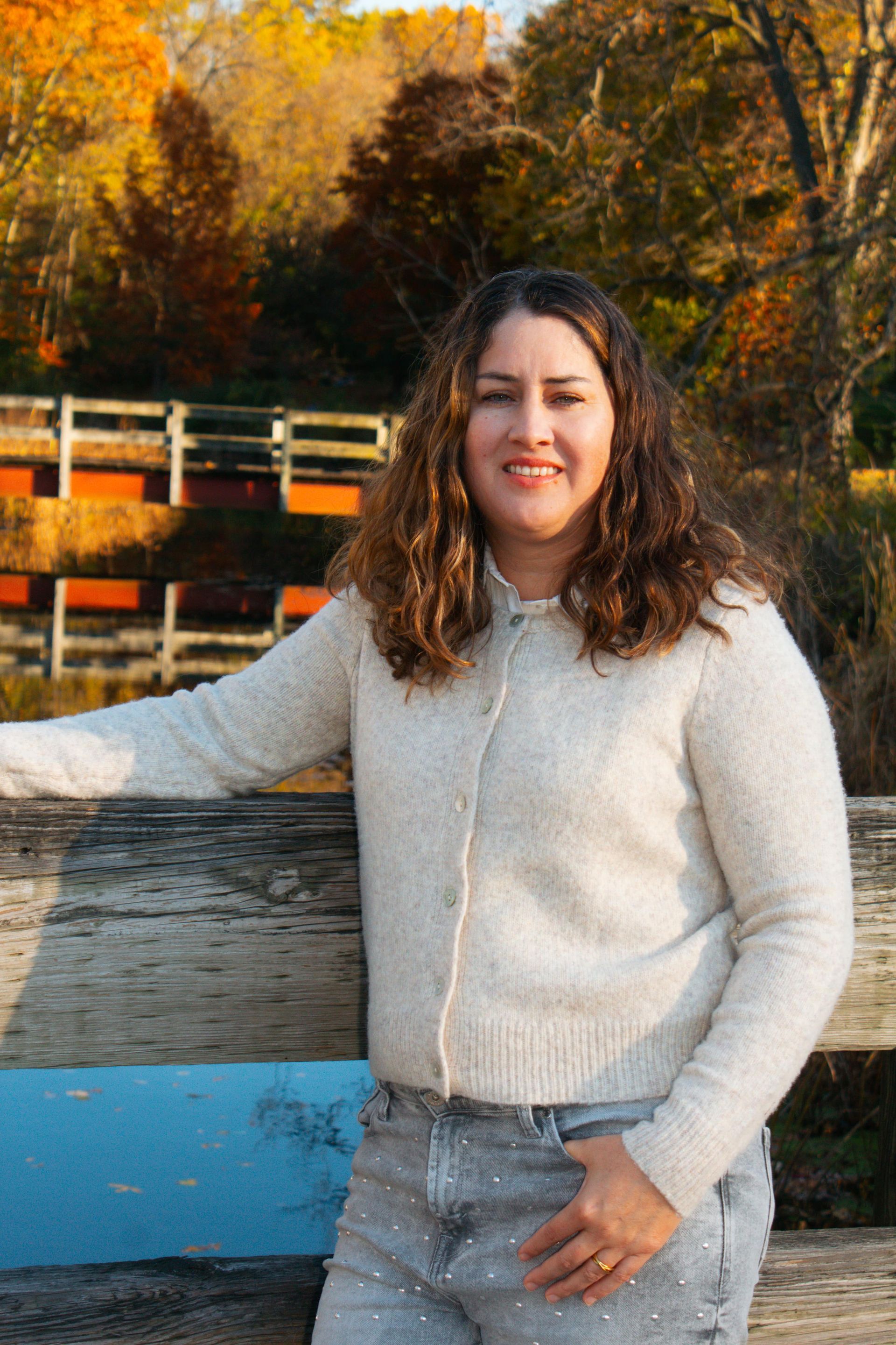 Woman in light sweater and embellished jeans leaning on a wooden structure, fall foliage in the background.