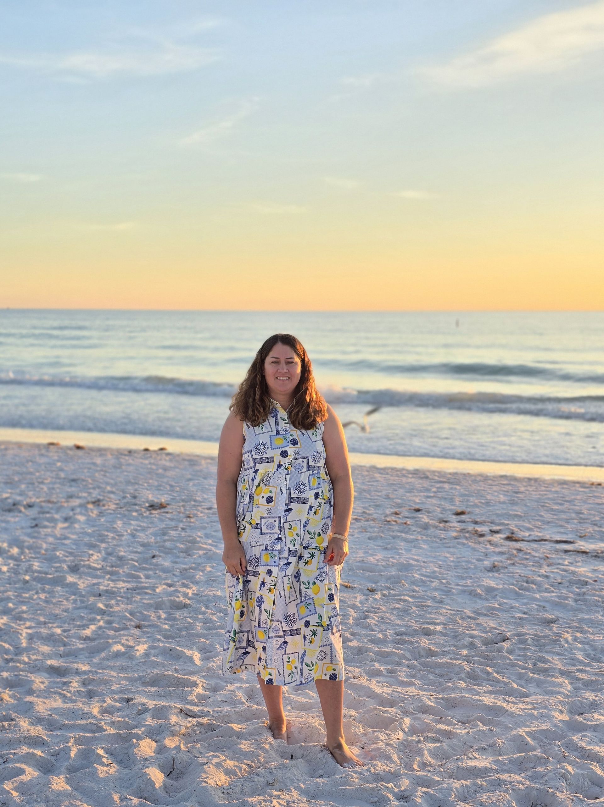 Woman in patterned dress stands on beach at sunset.