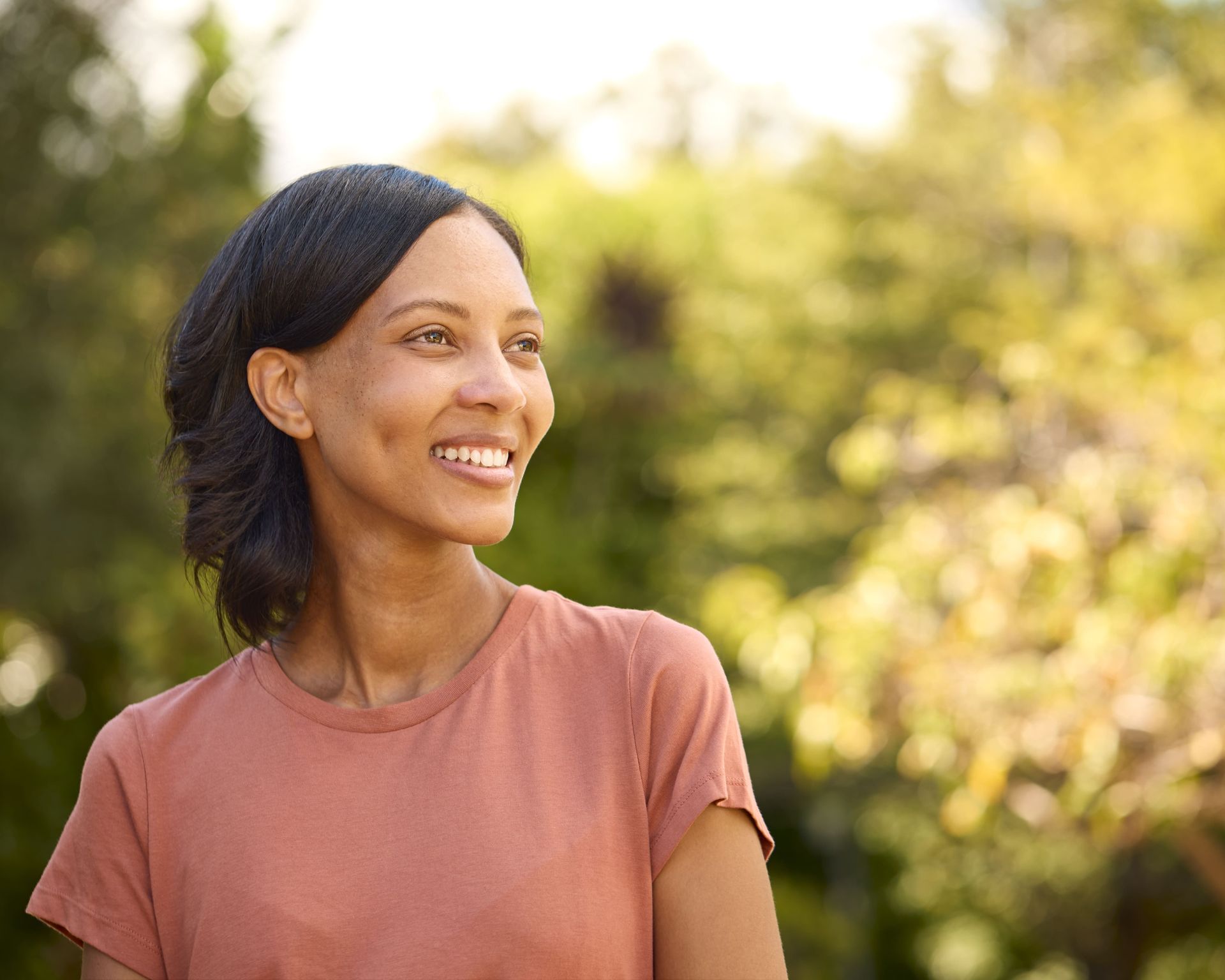 Woman with dark hair smiles, looking off to the side, wearing a pink shirt, outdoors.
