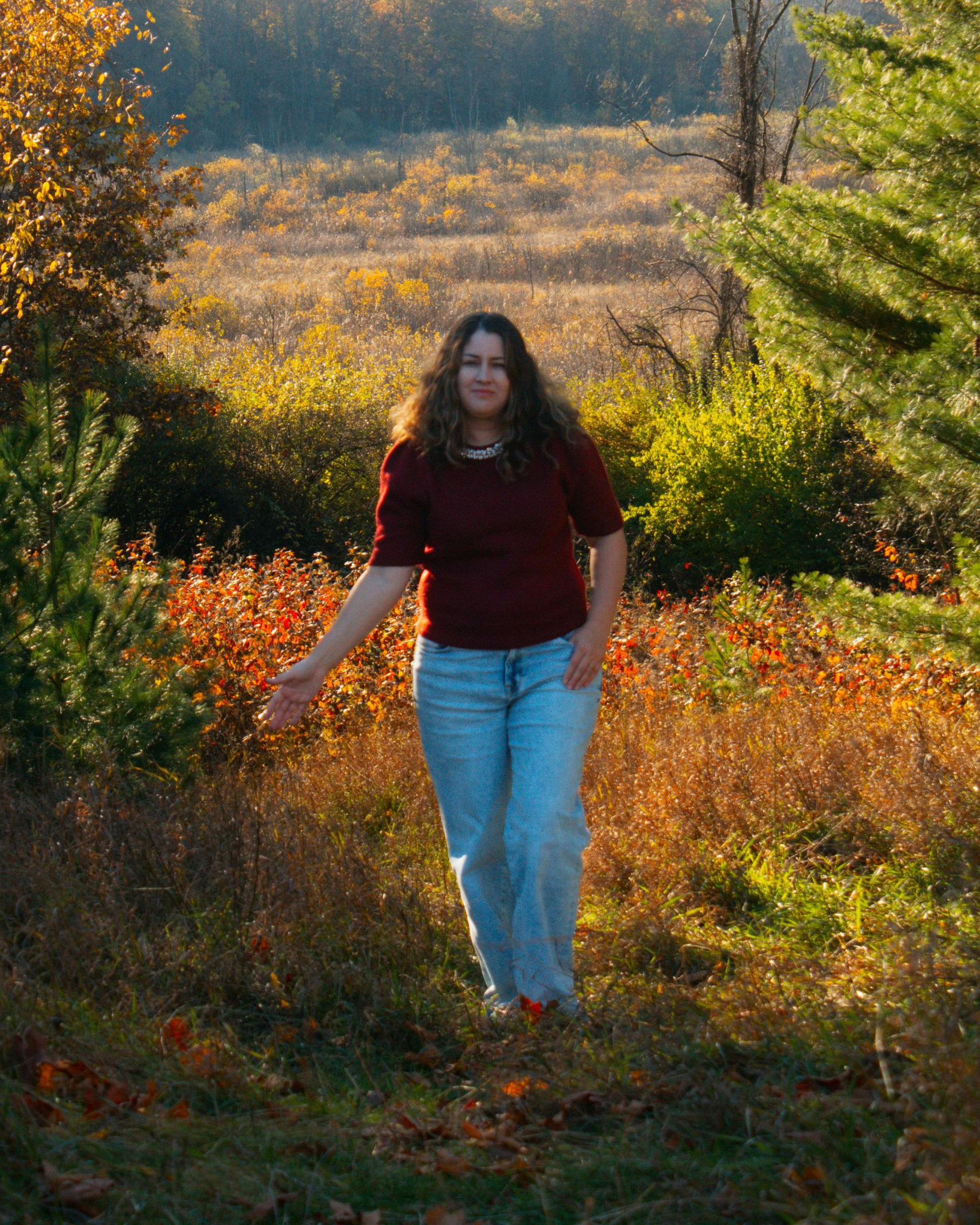Woman walks in field, reaching out. Autumn colors of red, orange, and green surround her.