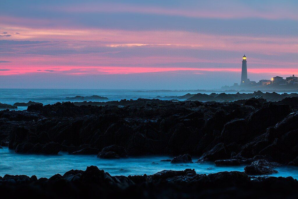 Atardecer rosa y azul sobre el océano con rocas oscuras en primer plano y un faro en el horizonte.