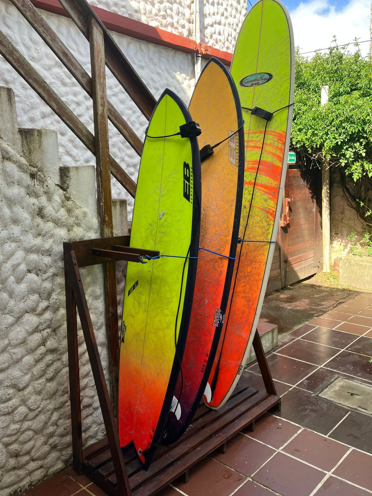 Vista exterior de un edificio blanco con detalles rojos, estructuras de madera y vegetación verde bajo un cielo azul.