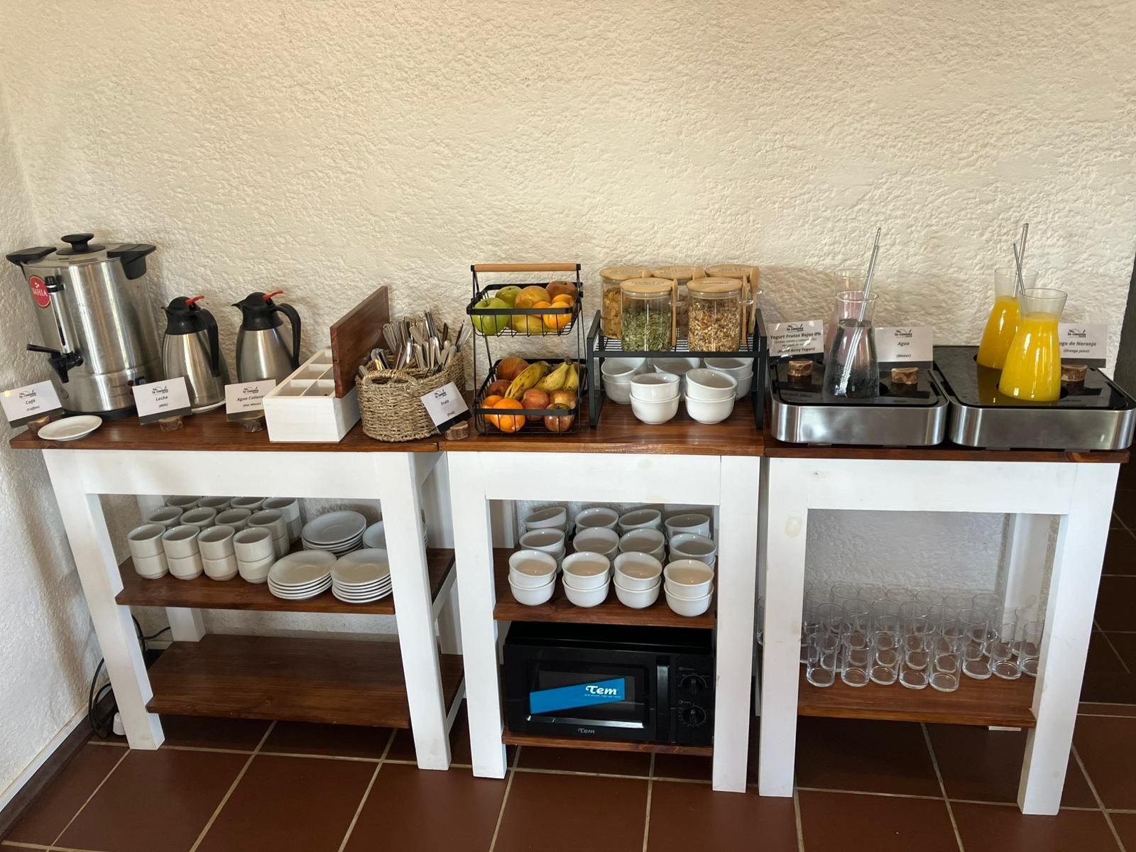 Desayuno buffet con café, té, fruta, cereales, zumo y una variedad de tazas y vasos sobre mesas blancas.
