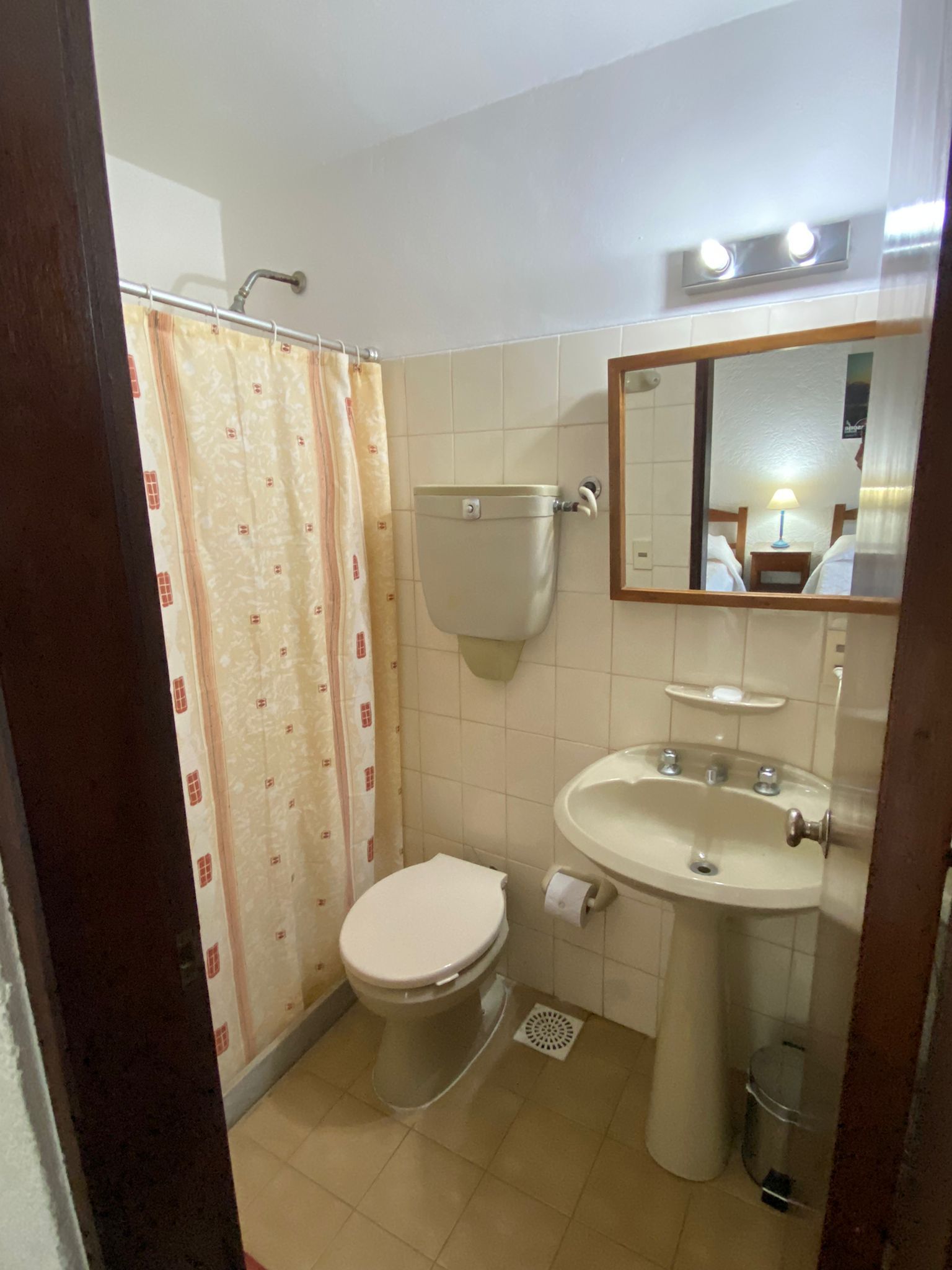 Baño pequeño con inodoro, lavabo, espejo y ducha con cortina. Paredes y suelo color beige.