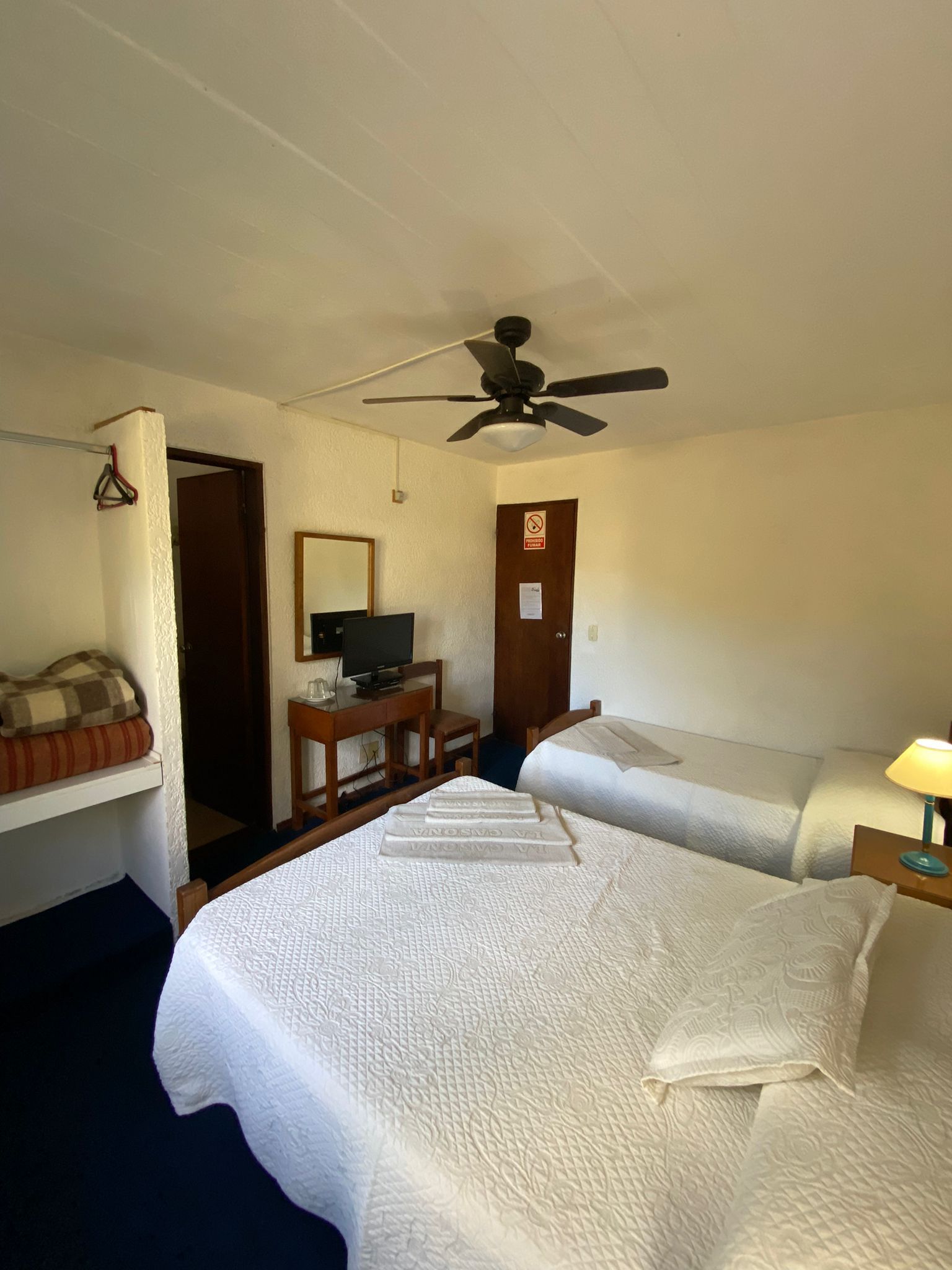 Dormitorio con dos camas, televisor, cómoda y ventilador de techo. Edredones blancos, alfombra azul, paredes beige.