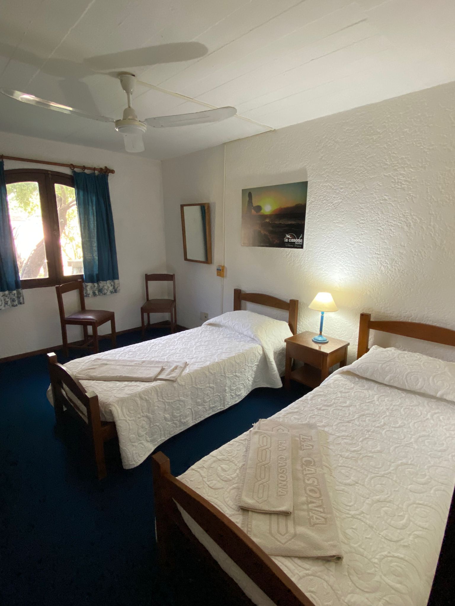 Dos camas en una habitación con ventana, sillas y una mesita de noche.