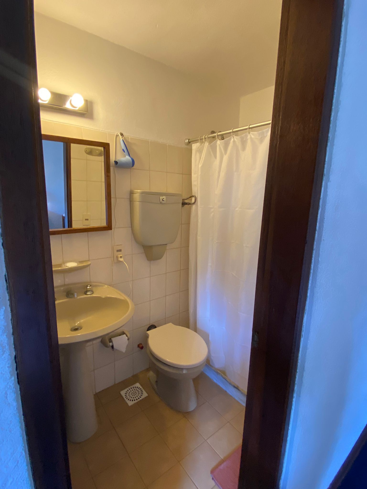 Baño pequeño con sanitarios blancos, cortina de ducha, paredes color beige y marco de puerta de madera.