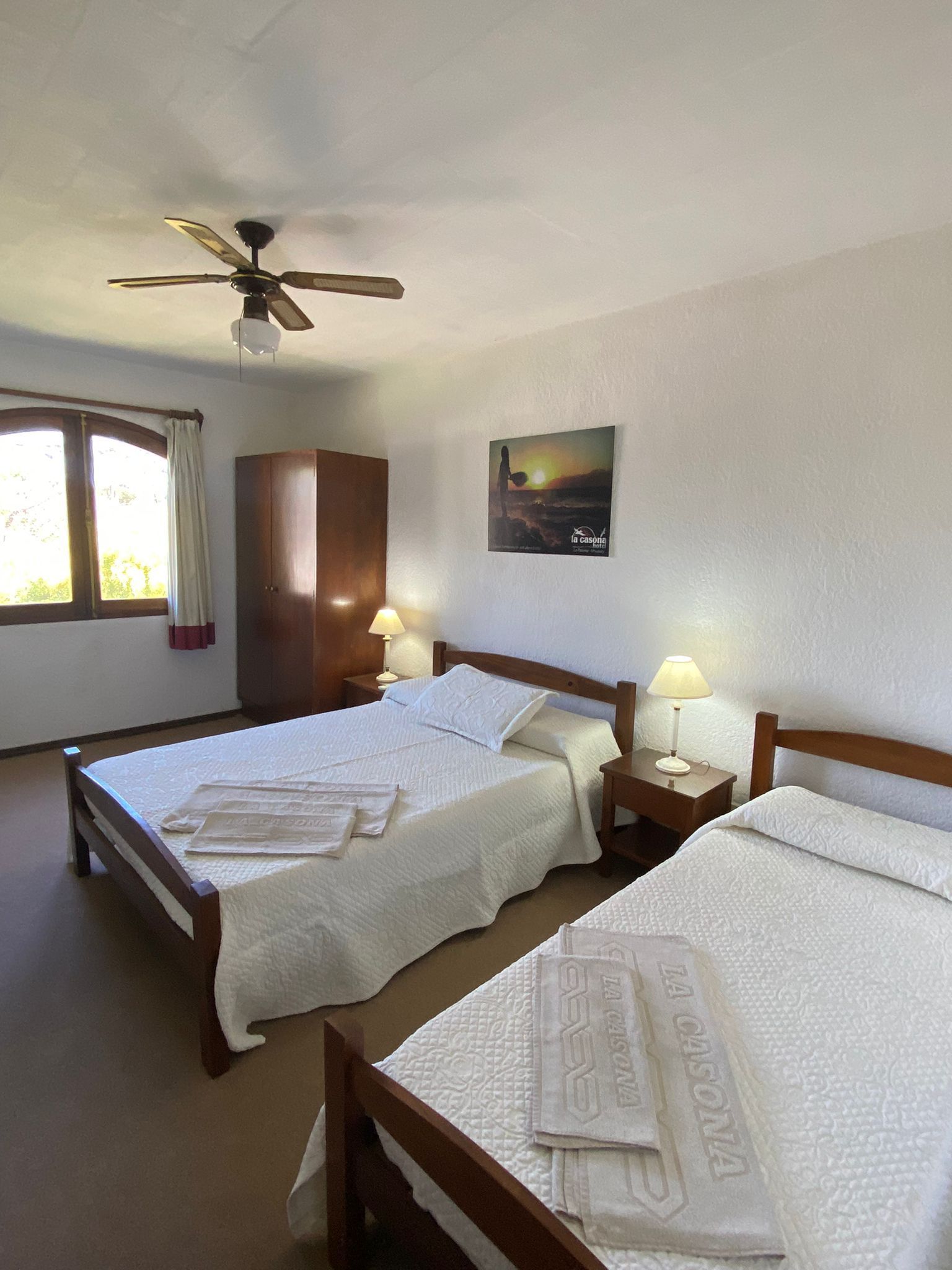 Dormitorio con dos camas, estructuras de madera, ropa de cama blanca. Ventana, ventilador de techo y armario.