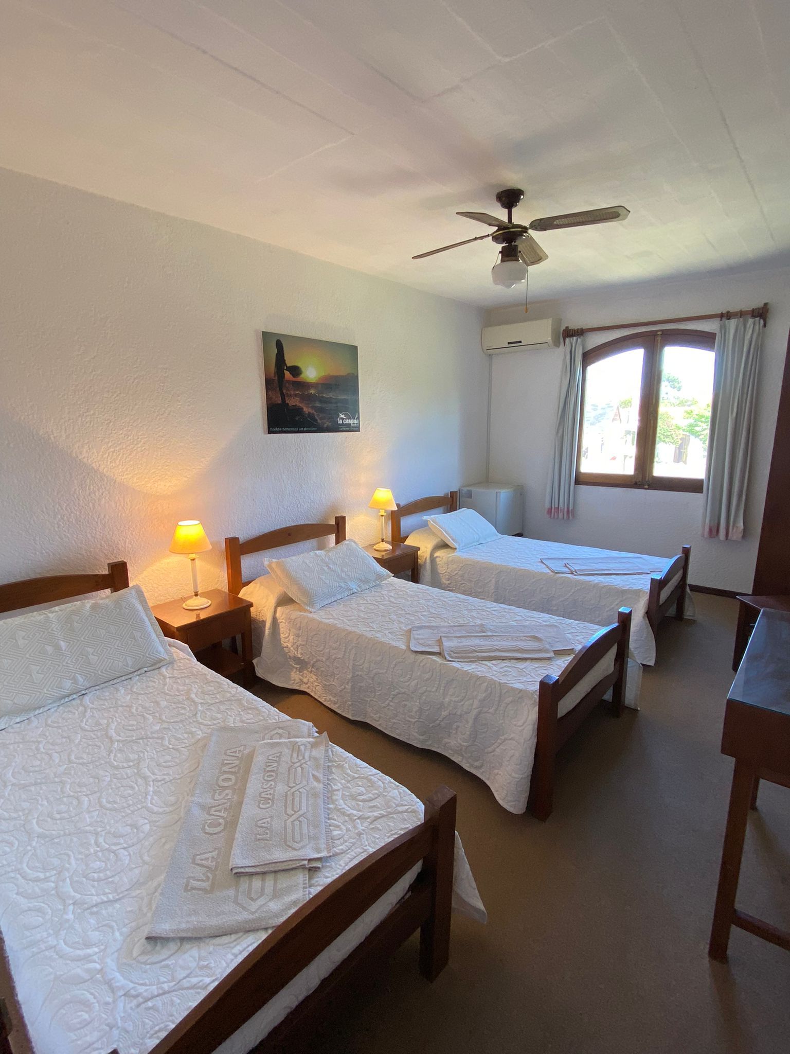 Tres camas en una habitación, dos con lámparas en las mesitas de noche. Ventana con cortinas, cuadro en la pared.