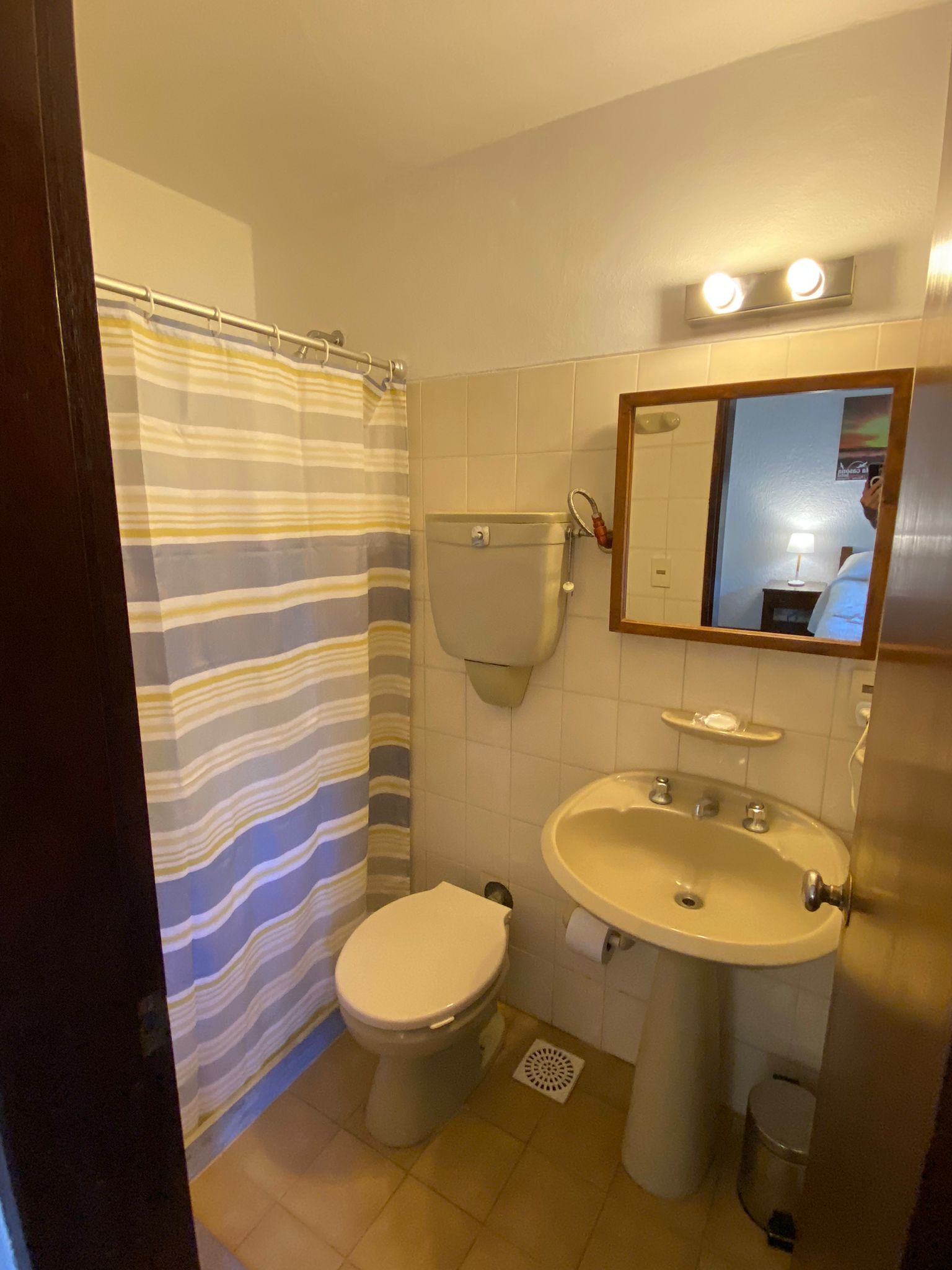 Baño con inodoro, lavabo y ducha. Suelo beige y paredes blancas. Cortina de ducha a rayas.