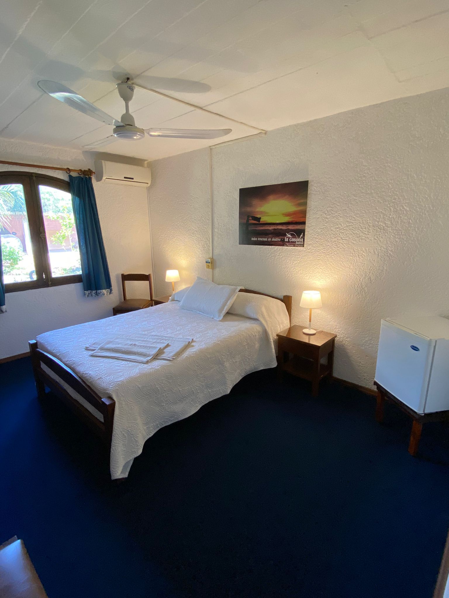 Dormitorio con dos camas, televisor, cómoda y ventilador de techo. Edredones blancos, alfombra azul, paredes beige.