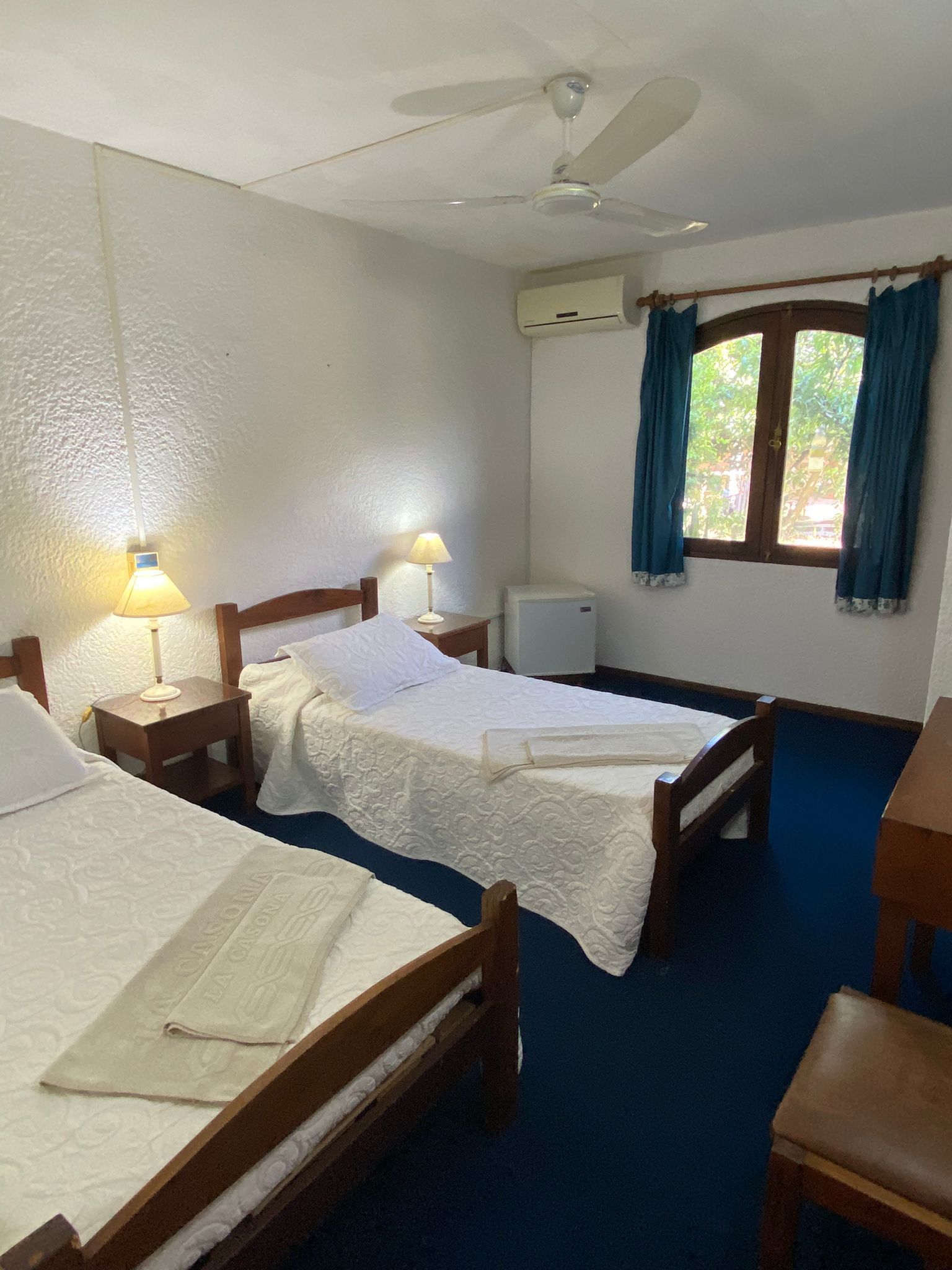 Dormitorio con dos camas, mesitas de noche con lámparas, ventana con cortinas azules y mininevera.