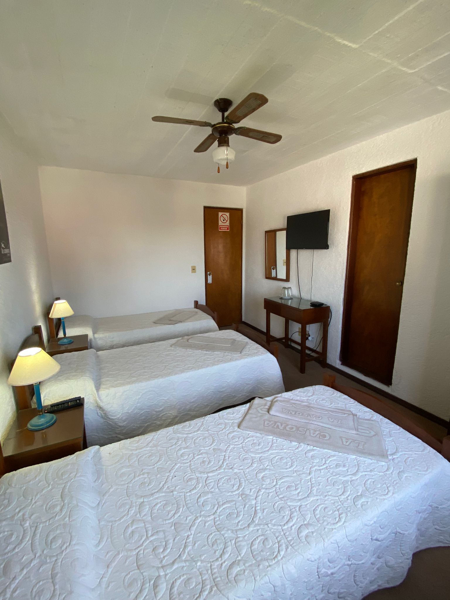 Habitación de hotel con tres camas, televisión, puerta de madera y ventilador de techo.