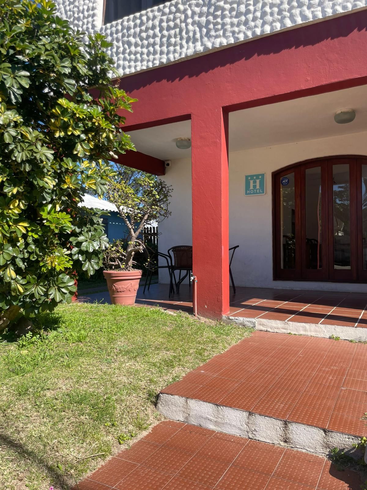 Exterior de un edificio con detalles en rojo, un porche cubierto y un pequeño patio con un árbol y césped.