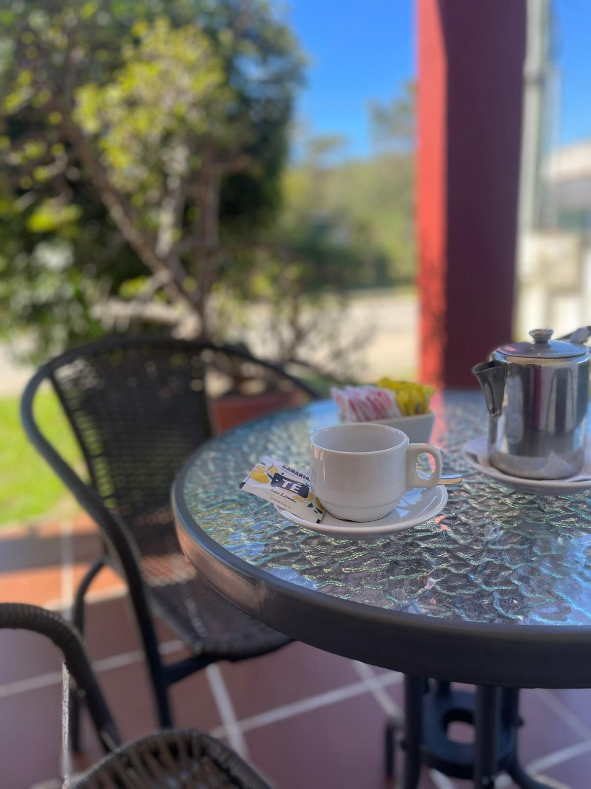 Mesa con taza de té, sobres de azúcar y tetera en un patio con vistas a los árboles.
