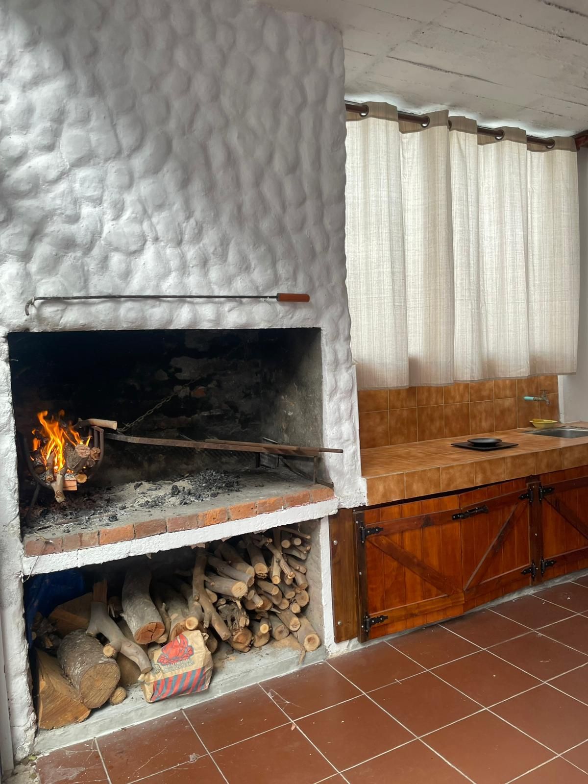 Chimenea de ladrillo con asador, leña y espacio para guardar leña; mostrador de madera adyacente.