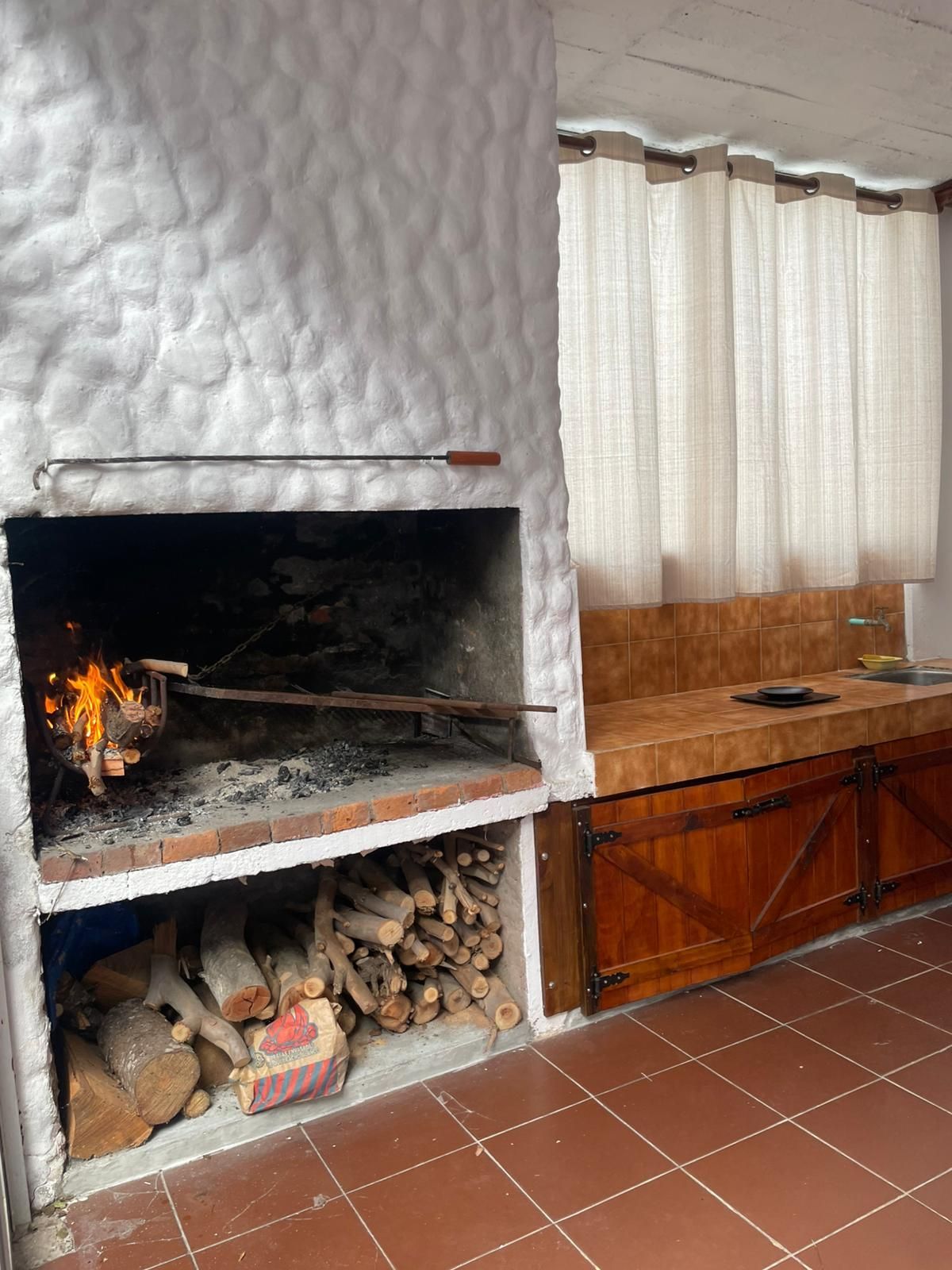 Chimenea de ladrillo con leña encendida, almacenamiento de leña y encimera de madera con cortinas.