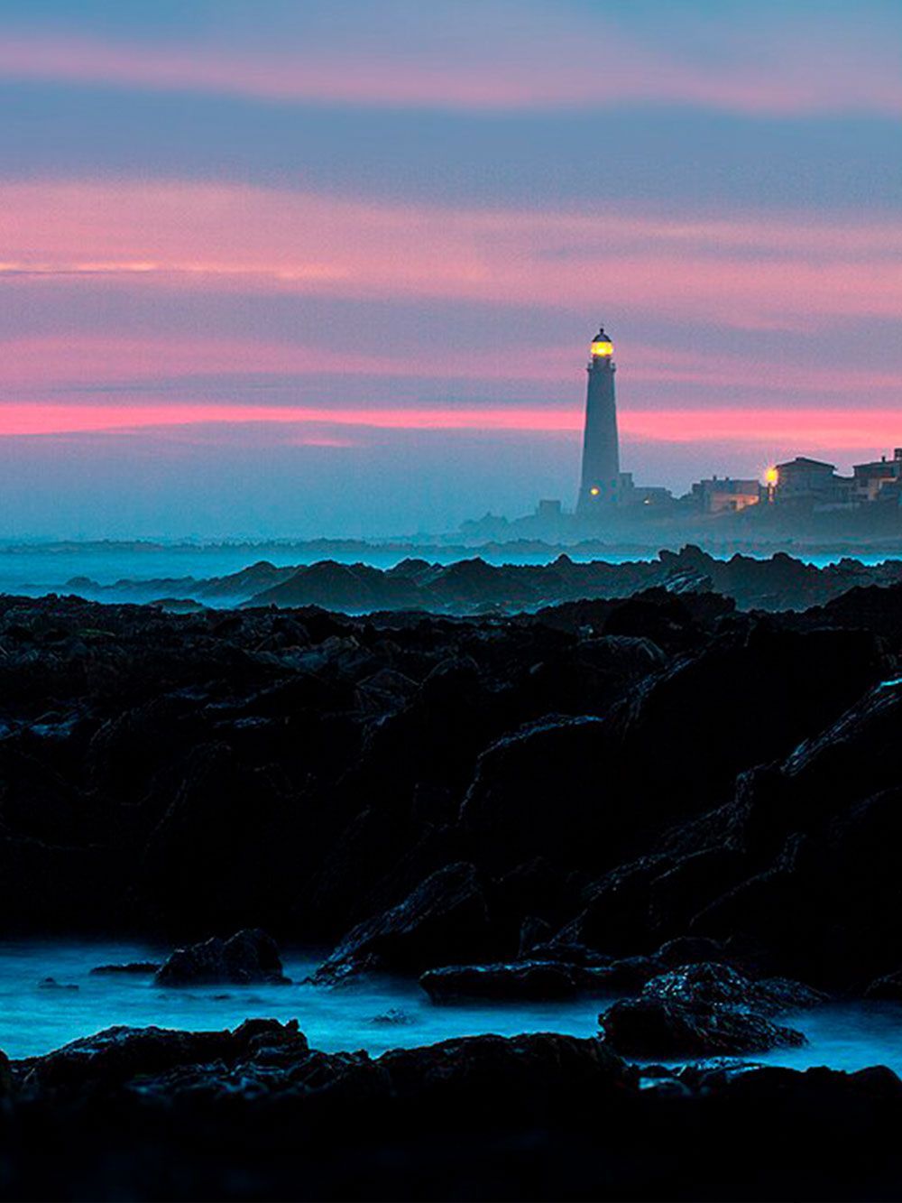 El faro se recorta contra un cielo de atardecer lleno de color. Rocas oscuras en primer plano, olas del océano.