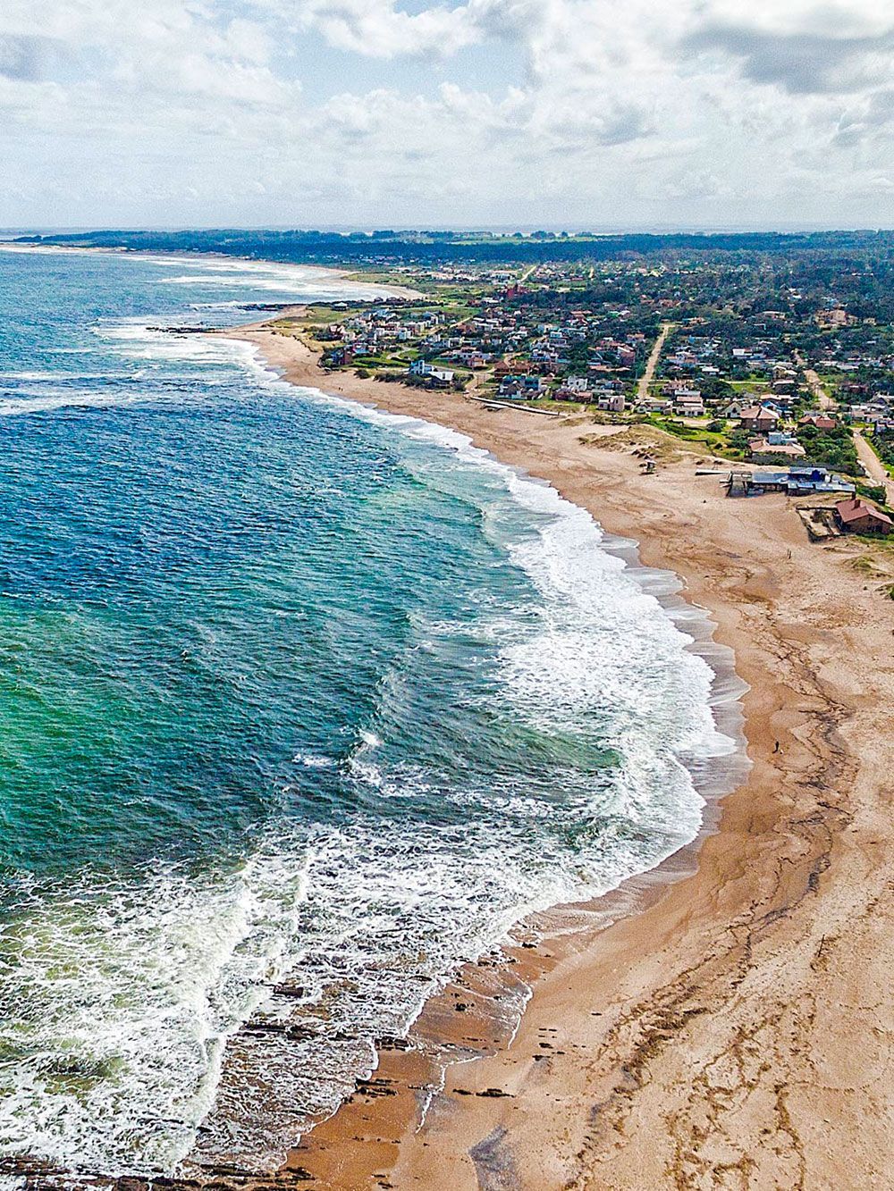 Vista aérea de una playa de arena y el océano, con olas rompiendo y un pueblo al fondo.