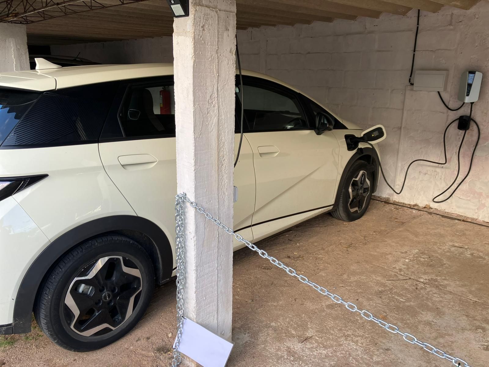Coche eléctrico blanco cargándose en un garaje. Cargador y cable a la derecha. Una cadena rodea un pilar.