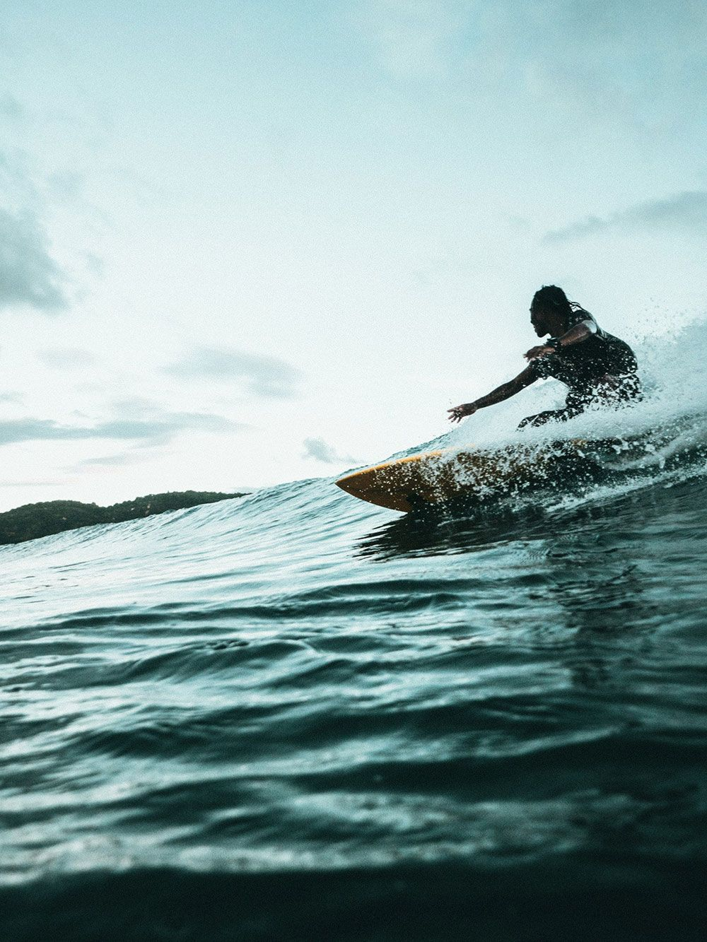Un surfista cabalga una ola con el brazo extendido. Al fondo se ve el agua del océano con un cielo nublado.