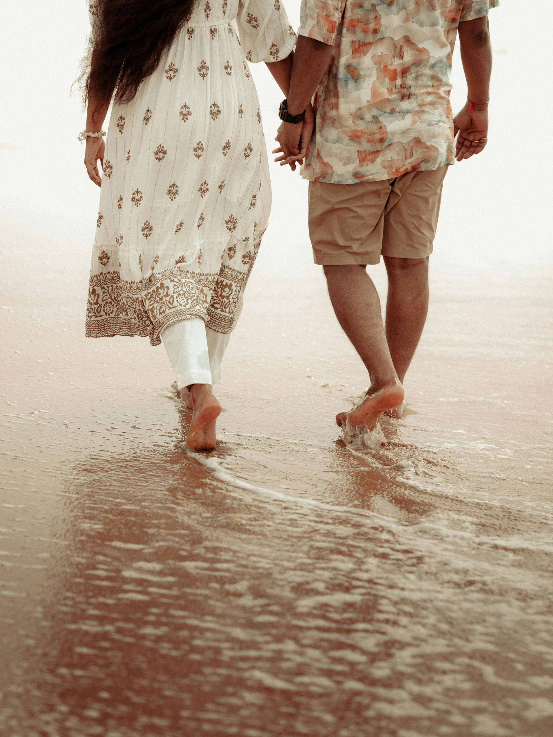 Pareja cogida de la mano, caminando descalzas por la playa, las olas les mojan los pies.