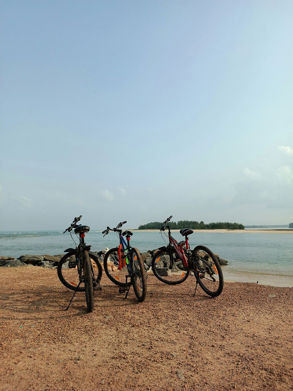 Tres bicicletas aparcadas en una playa de arena junto al océano, con una isla al fondo bajo un cielo azul.