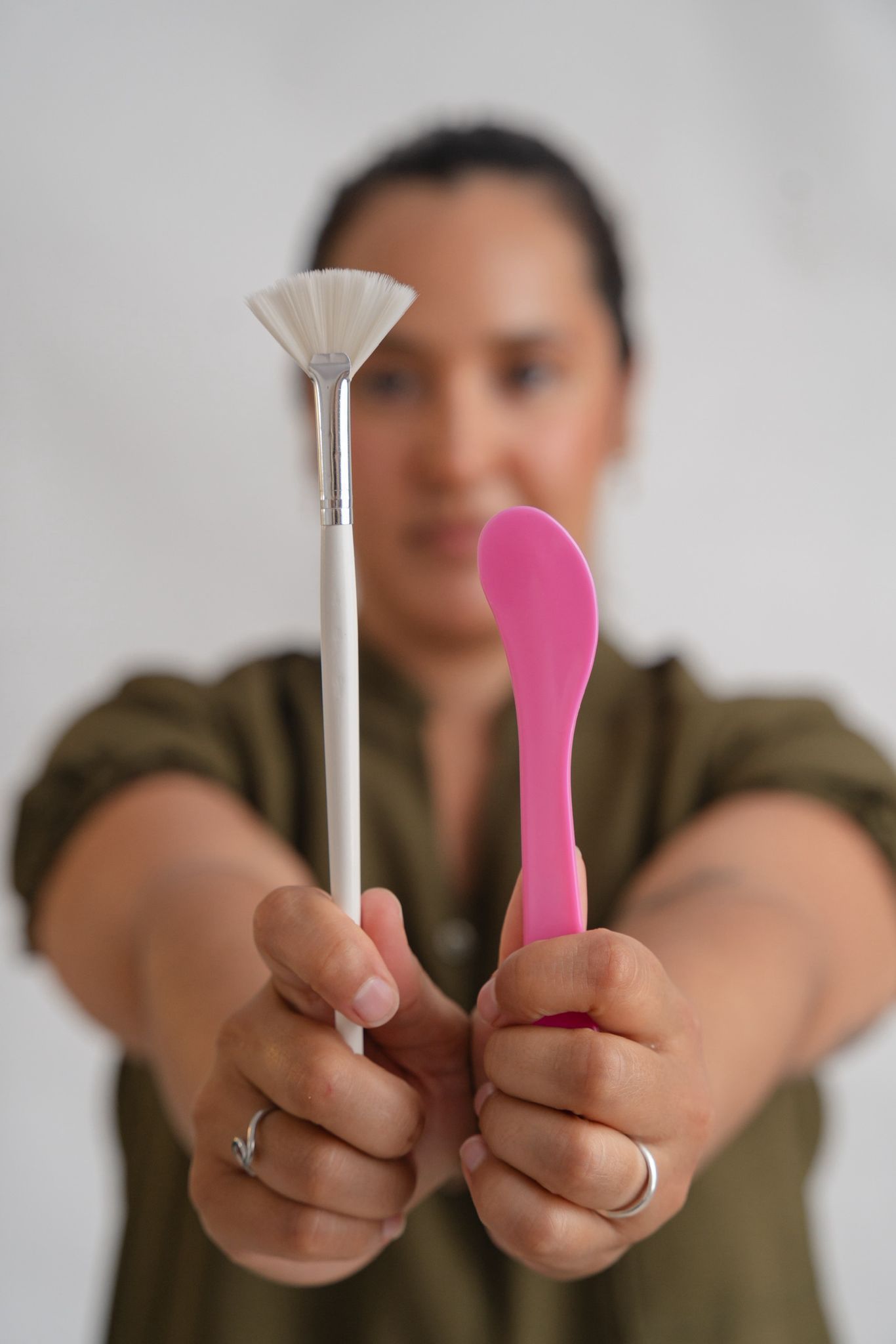 Mujer sosteniendo un pincel abanico y una espátula rosa, centrada, con un fondo borroso.