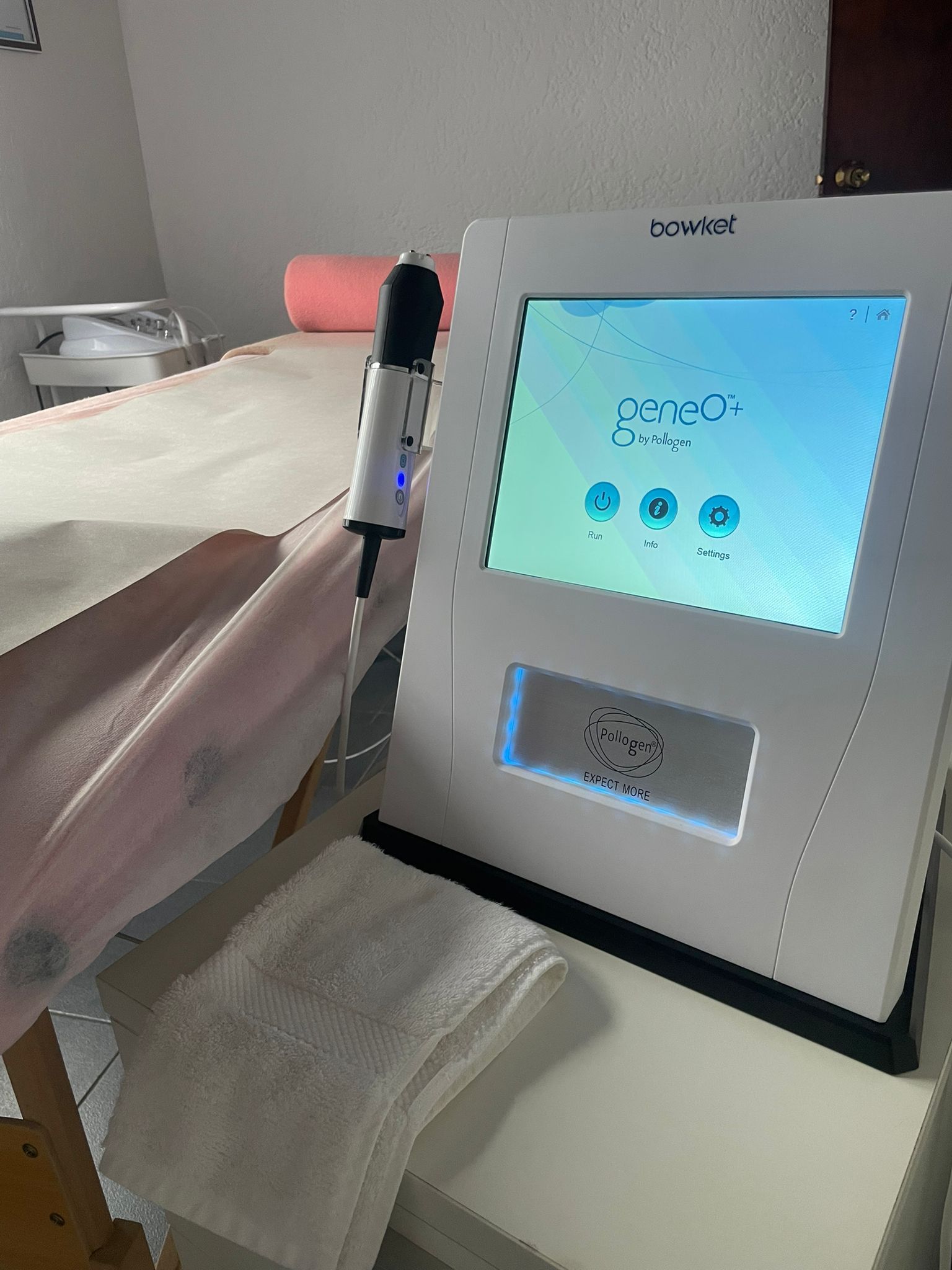 Dispositivo médico blanco con pantalla, sobre una mesa en una sala de tratamiento, al lado de una cama.