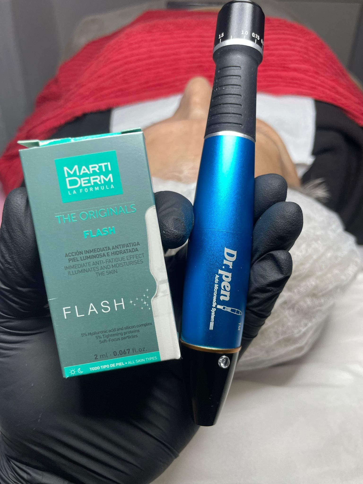 Mano sosteniendo un dispositivo Dr. Pen azul y una caja de ampollas Marti Derm Flash, con una persona en el fondo.