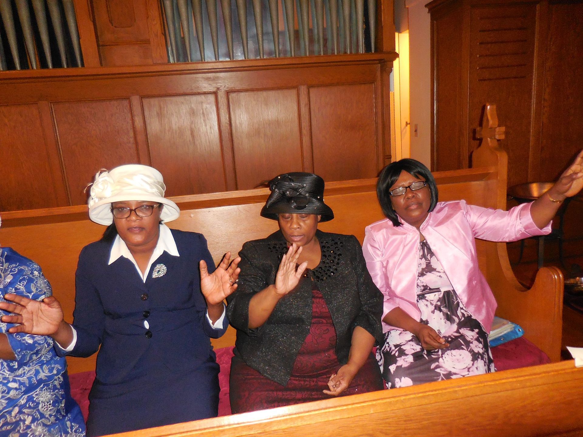 Three people sit in wooden pews with hands raised, wearing hats and formal attire inside a church sanctuary.