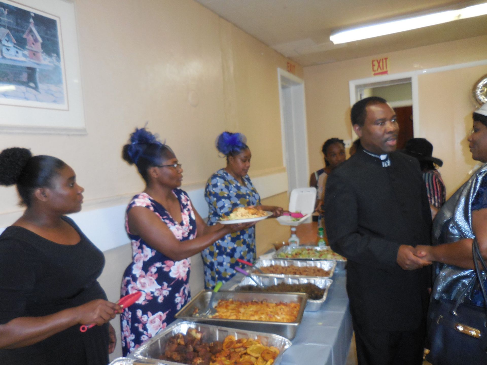 People serve buffet-style food at a gathering while a man in a black clerical shirt speaks with a guest.