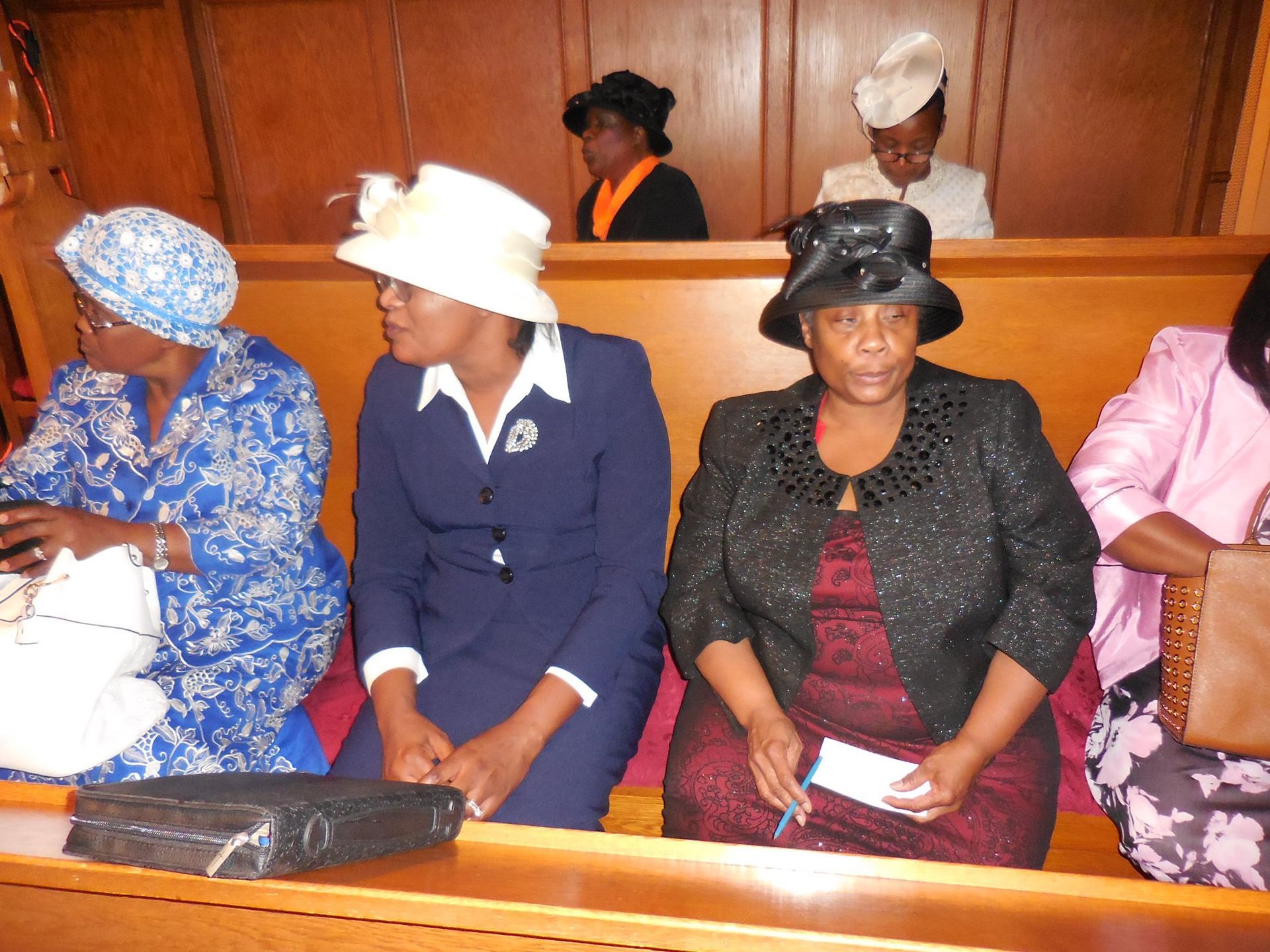 A row of people wearing hats and formal attire sit together on wooden church pews.