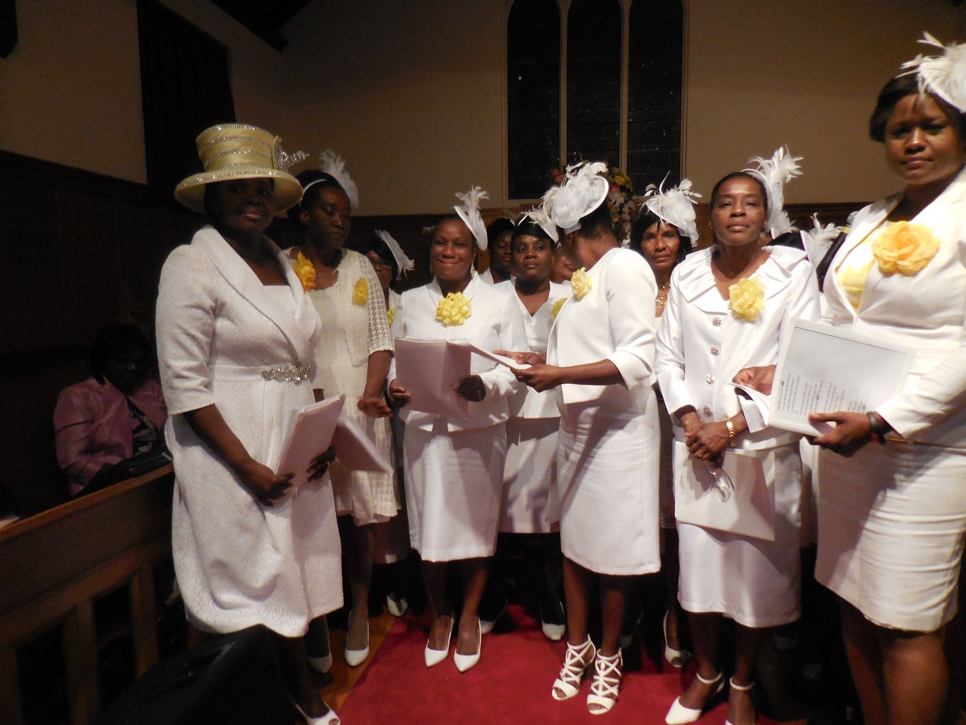 A group of people dressed in white suits and hats stand in a church, holding songbooks while singing together.