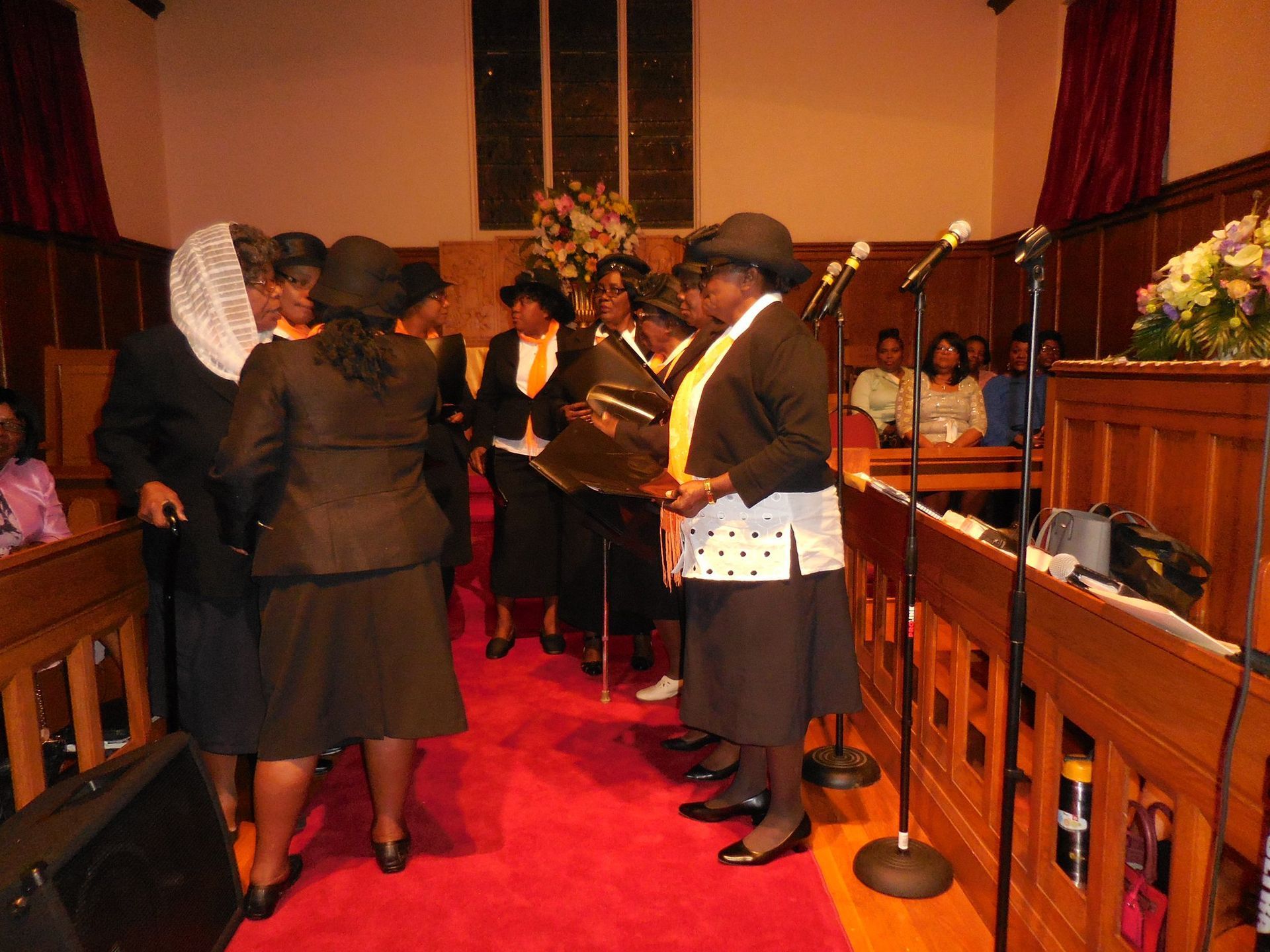 A group of people wearing hats and dark formal attire stand on a red carpet in a wood-paneled church sanctuary.