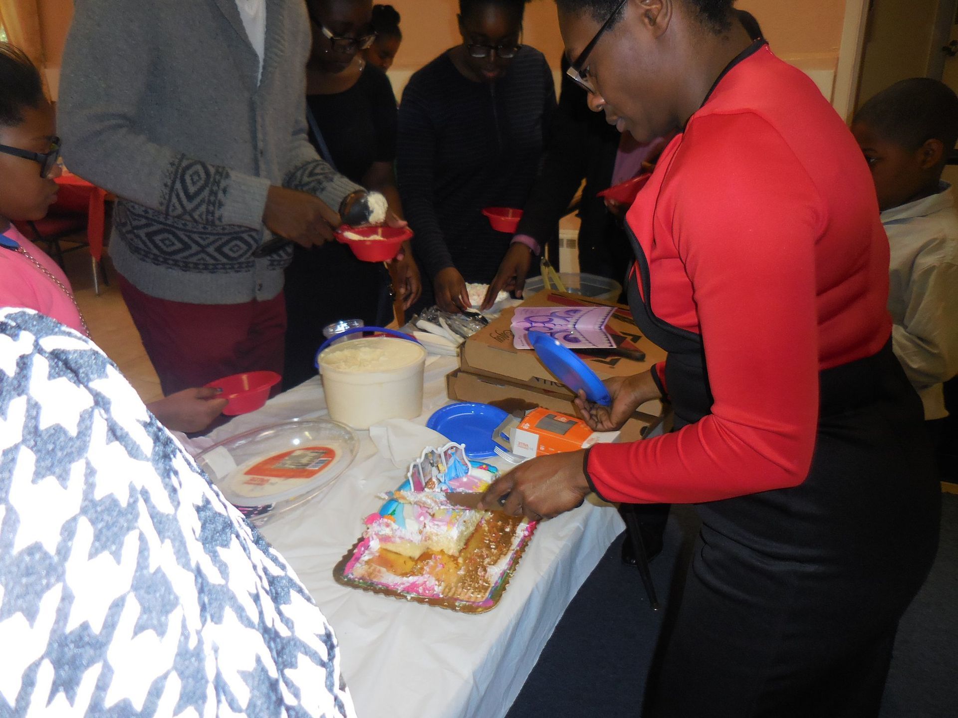 People gather around a table to cut and serve a decorated cake at an indoor event.