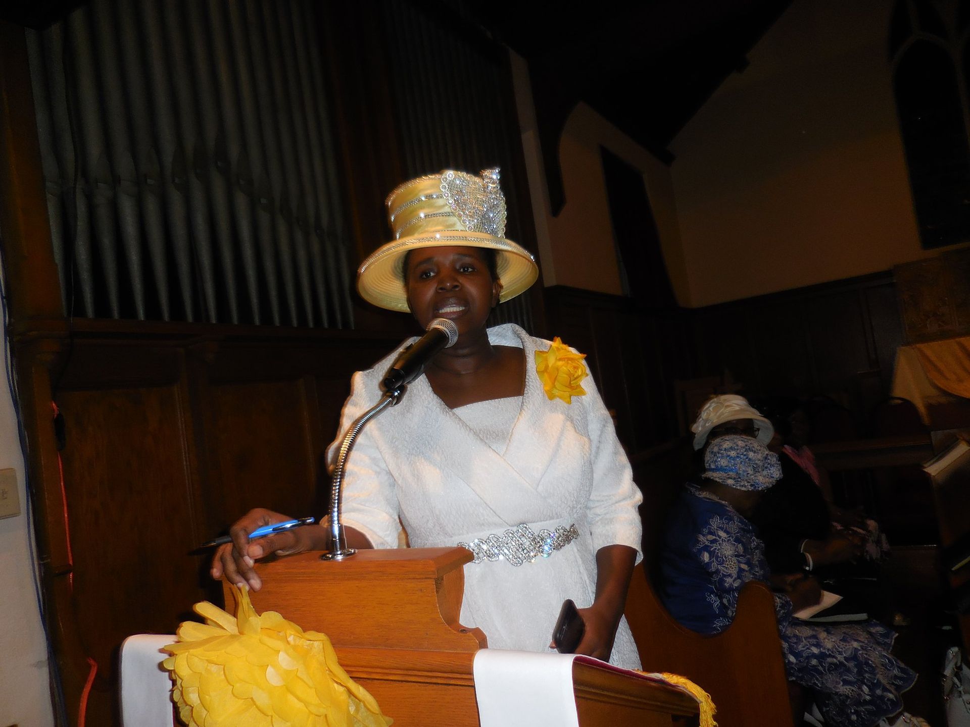 A person in a cream hat and white dress speaks at a wooden podium in a church setting.