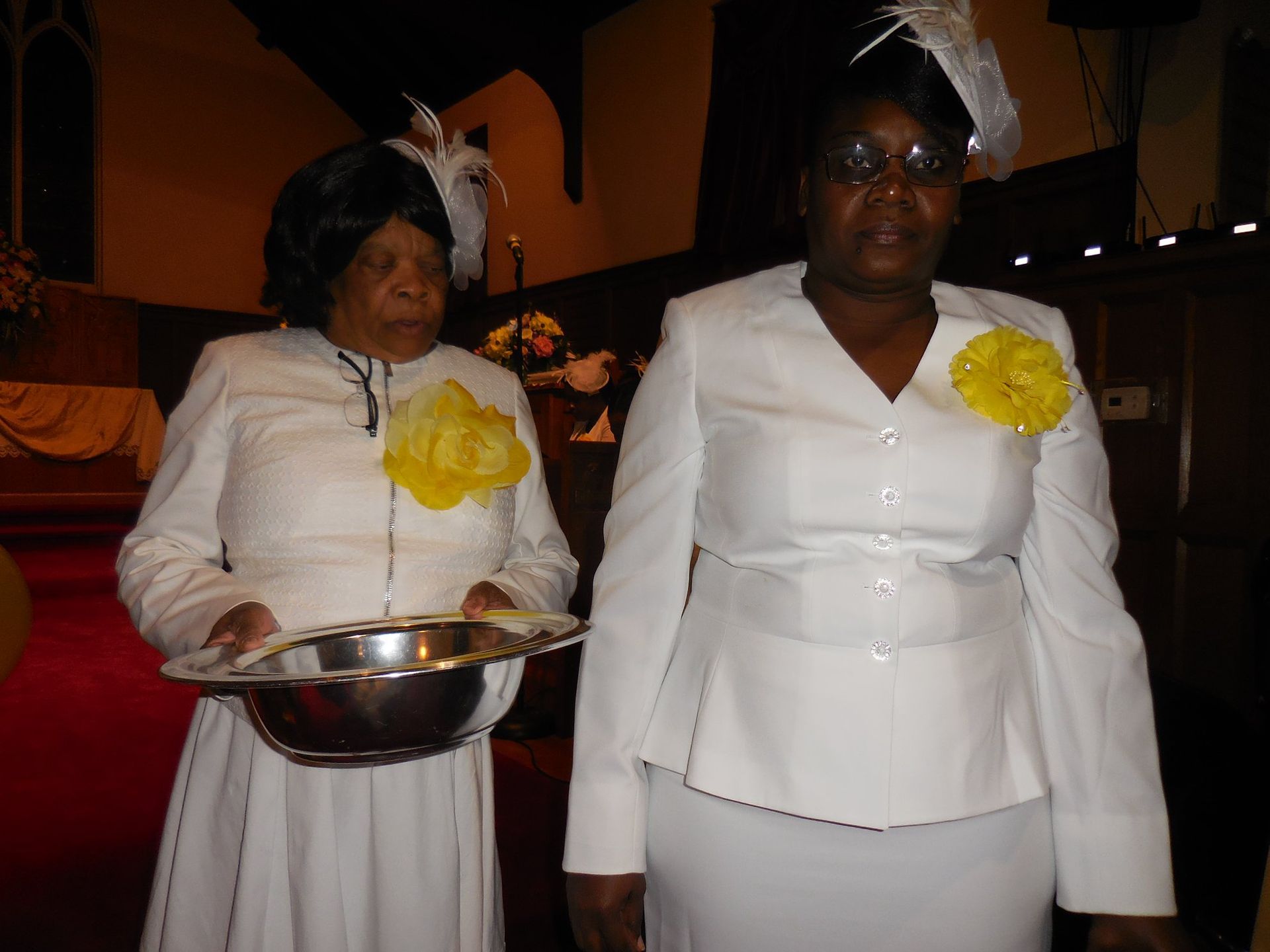 Two people in formal white attire and hats with yellow corsages stand indoors, one holding a silver basin.