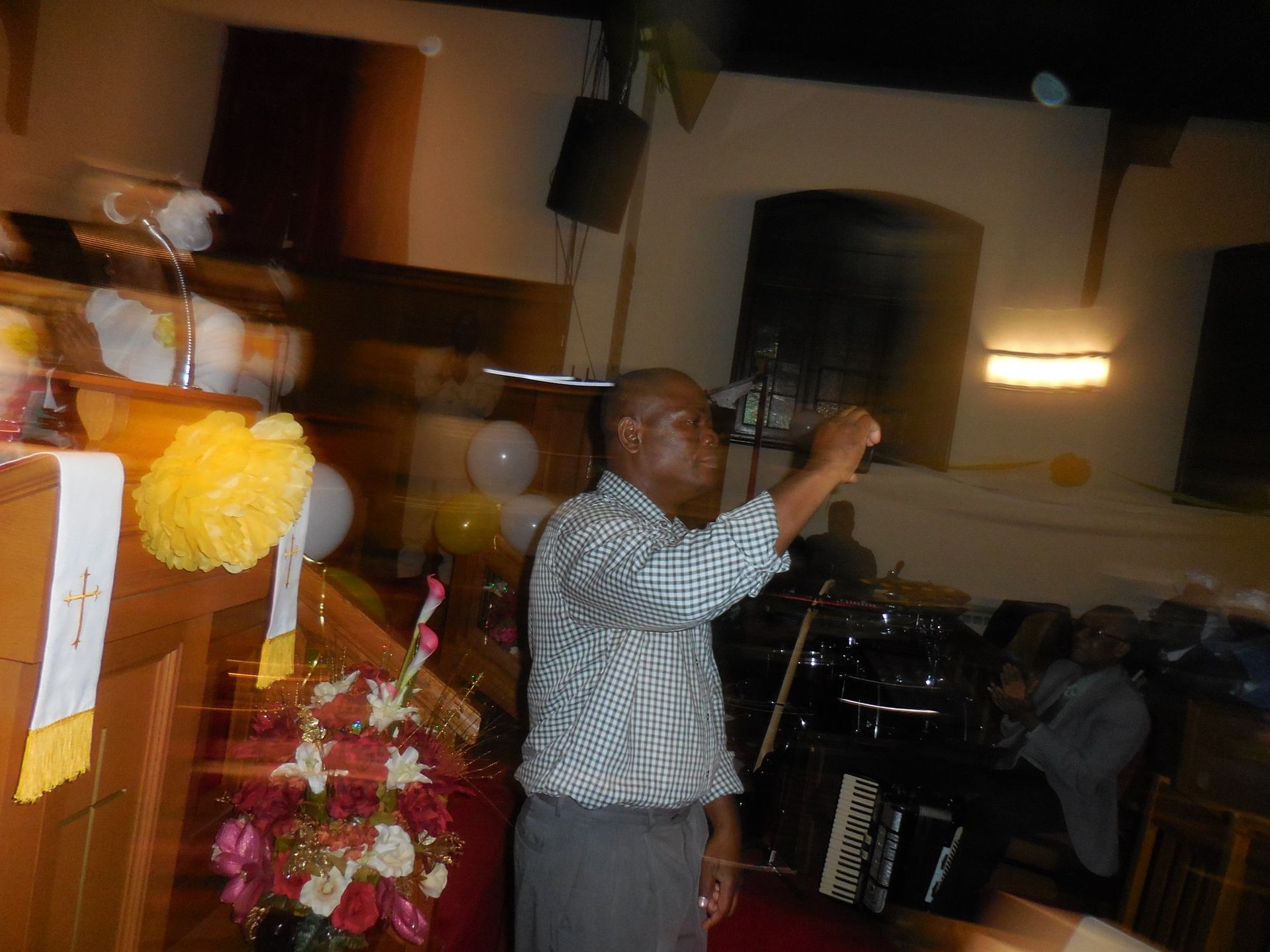 A person in a checkered shirt gestures while speaking from a lectern in a dimly lit room decorated with yellow flowers.