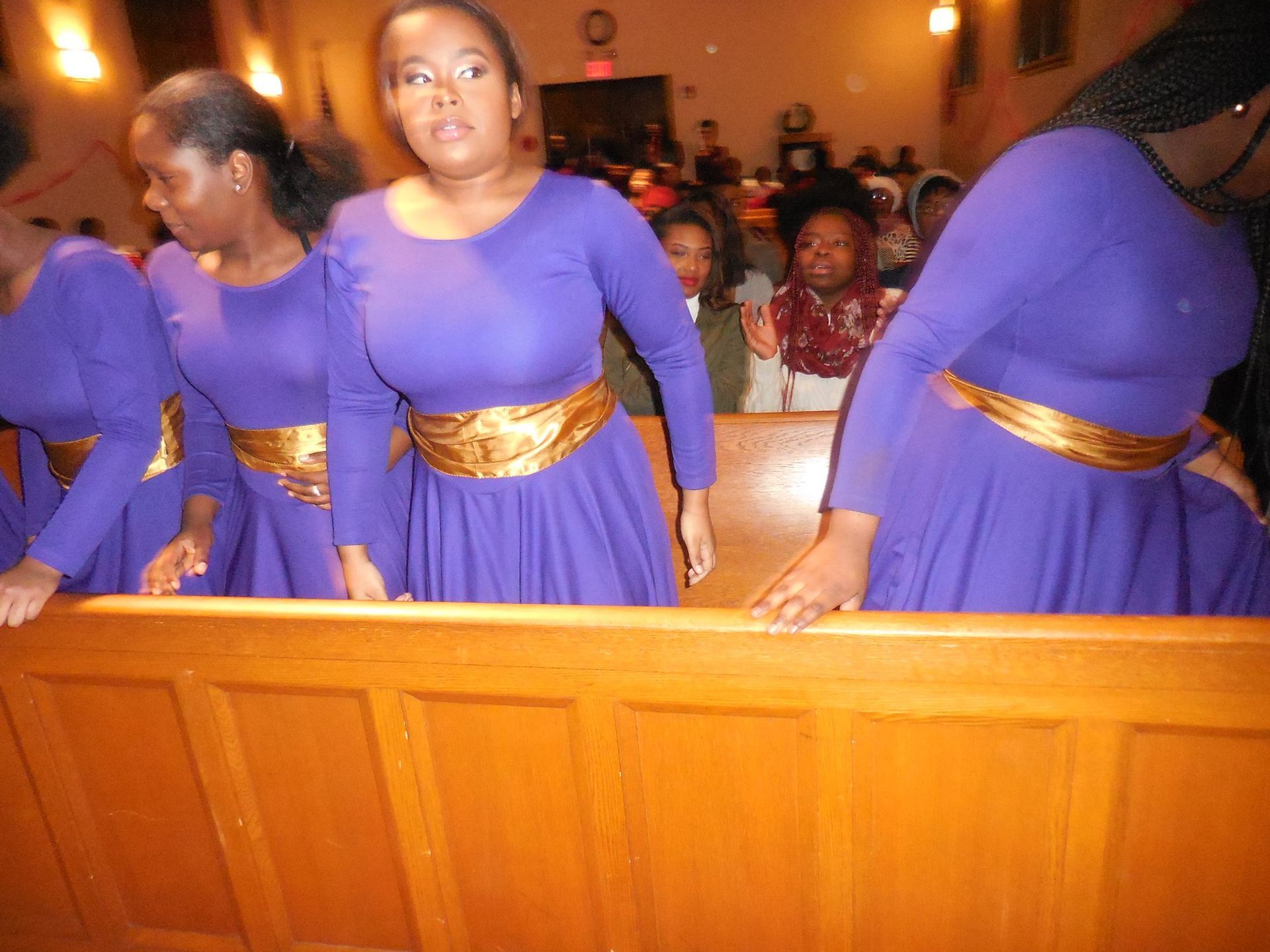 People wearing matching purple dresses with gold belts standing in a wooden pew during a church service.