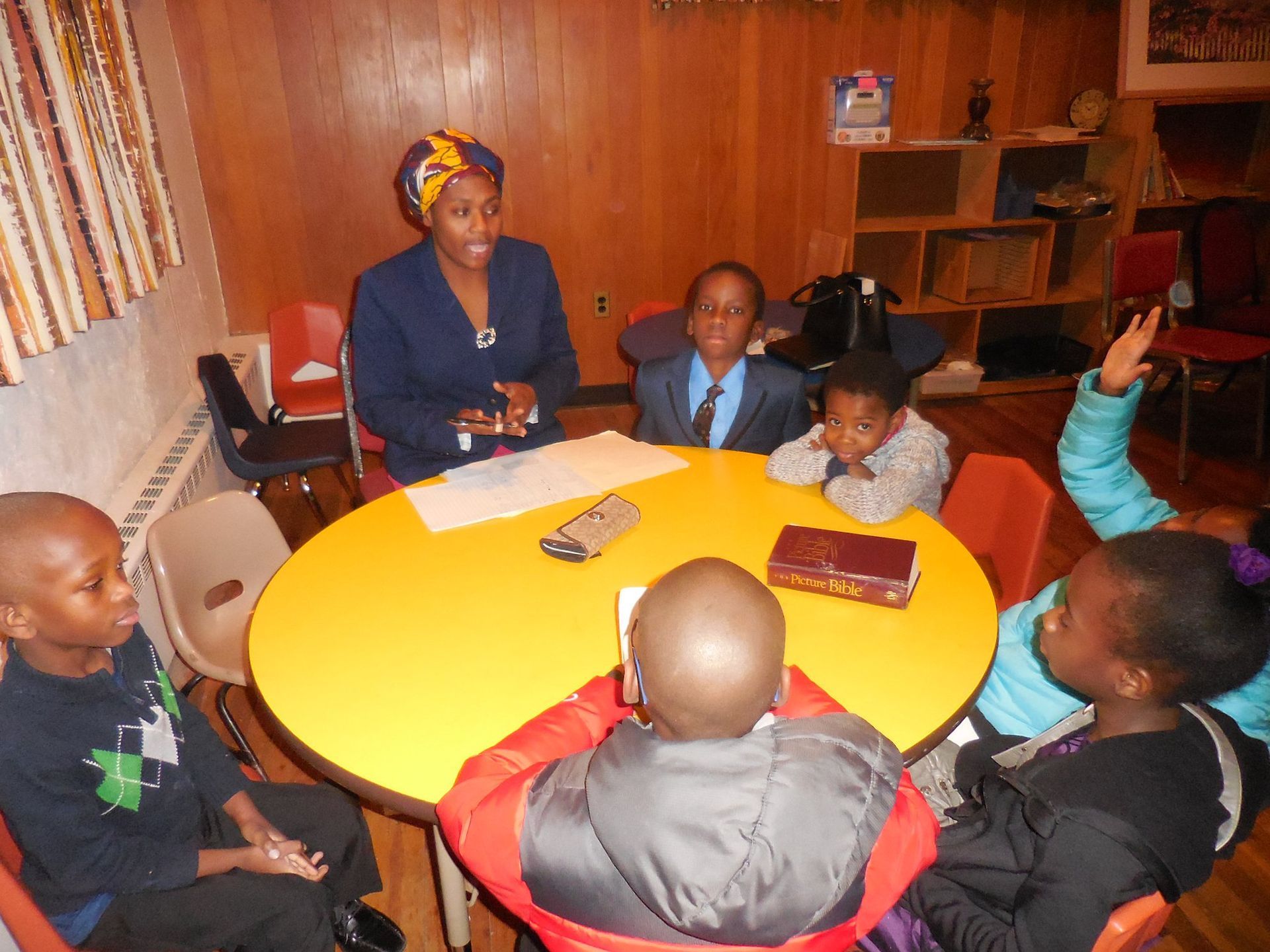 A small group sits around a yellow circular table with an adult leader, one student raising their hand in a classroom.