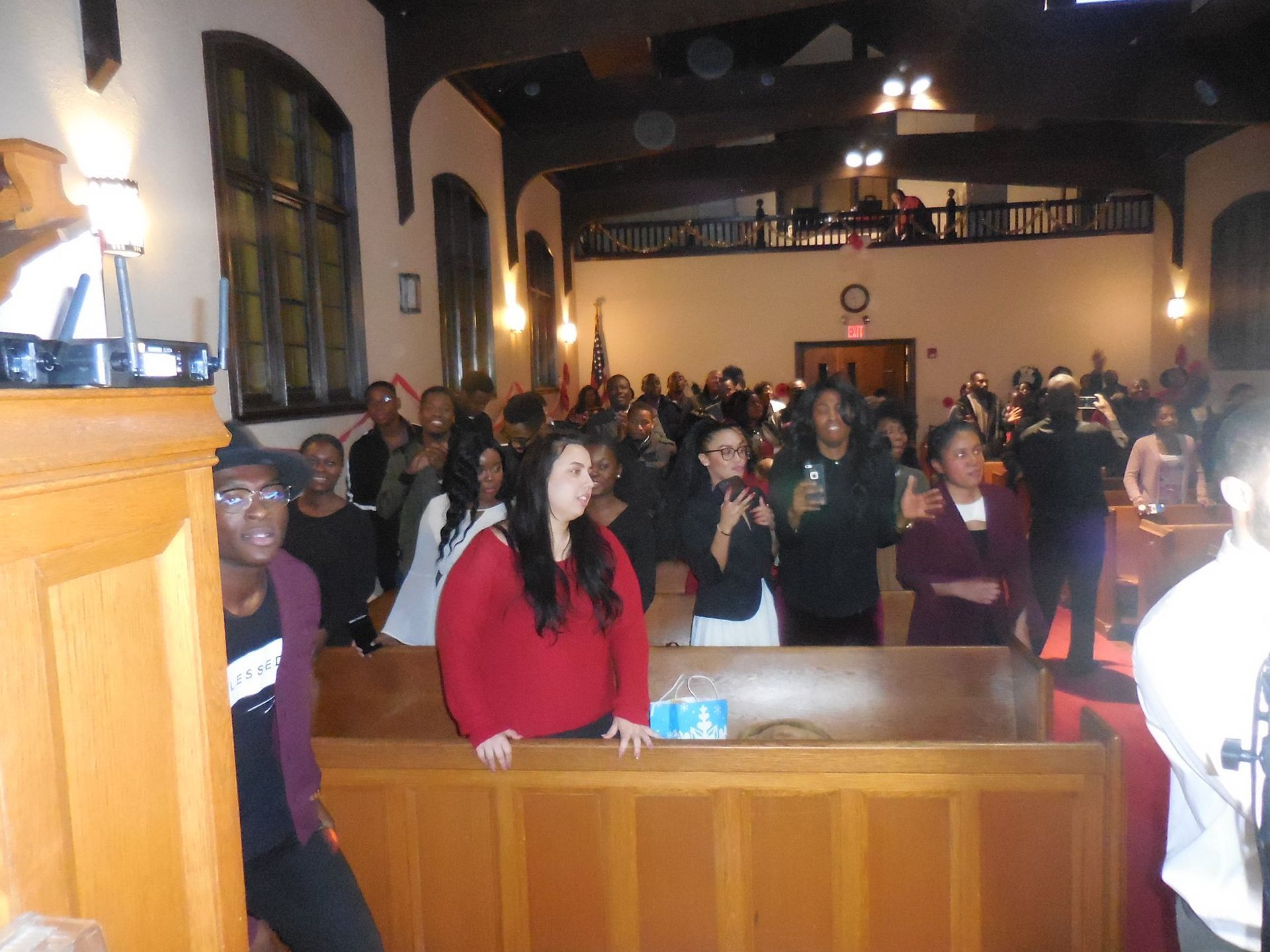People stand and look toward the front during a service inside a church with wooden pews and arched windows.