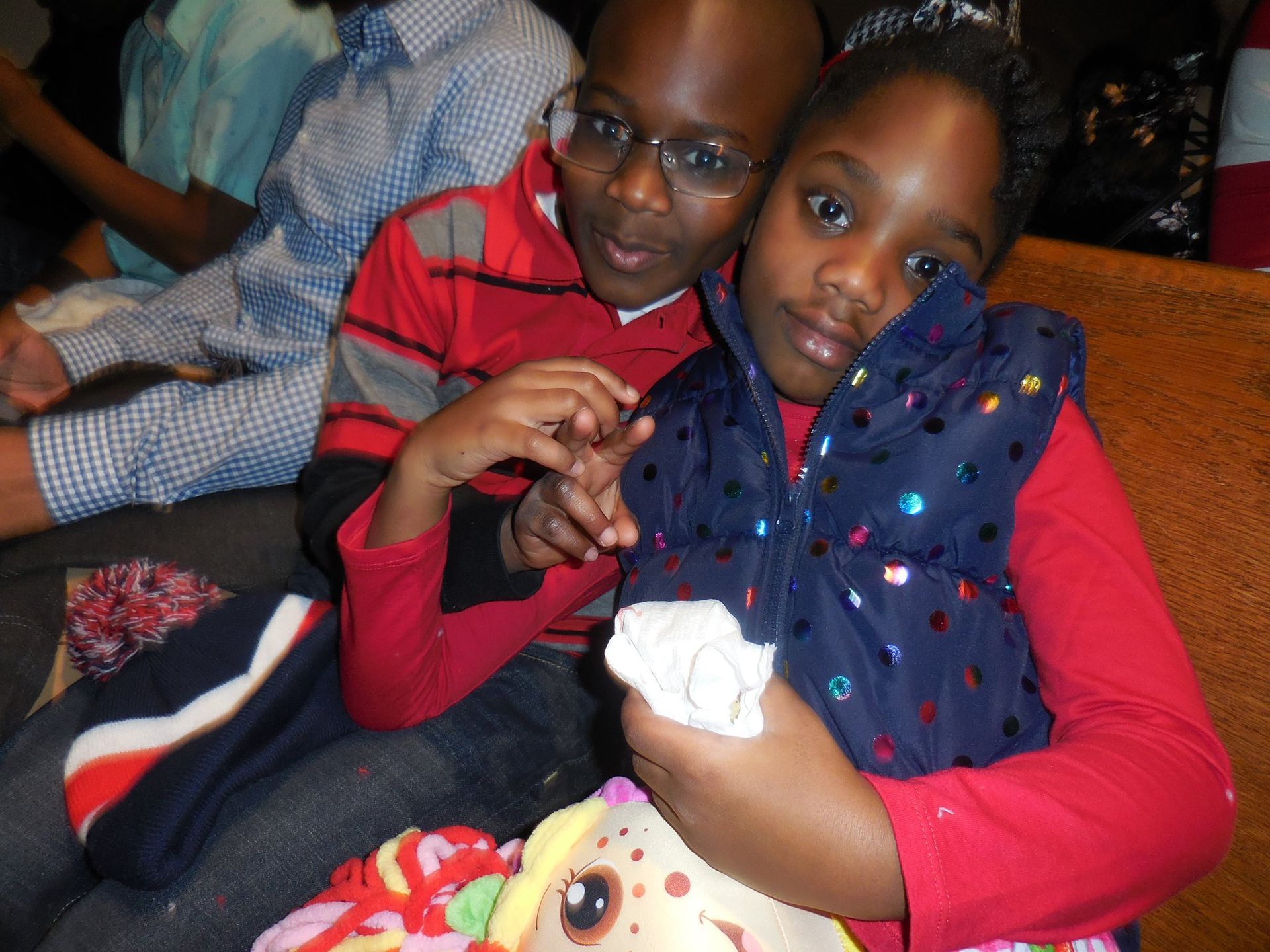Two children sit together on a bench; one wears a red striped sweater, the other wears a blue glittery vest.
