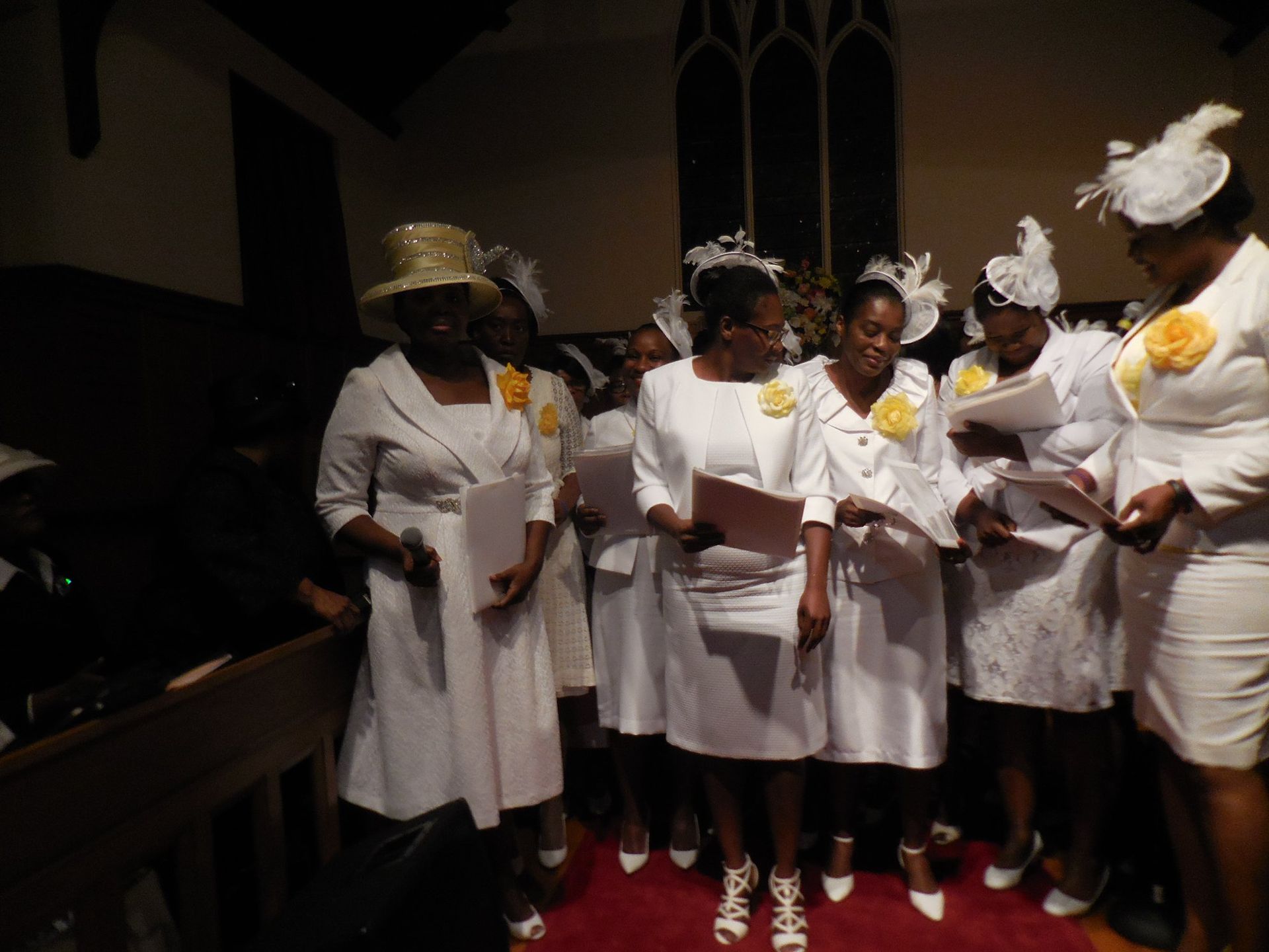 A group of people wearing white dresses and decorative hats stand in a church, holding papers while singing or reading.