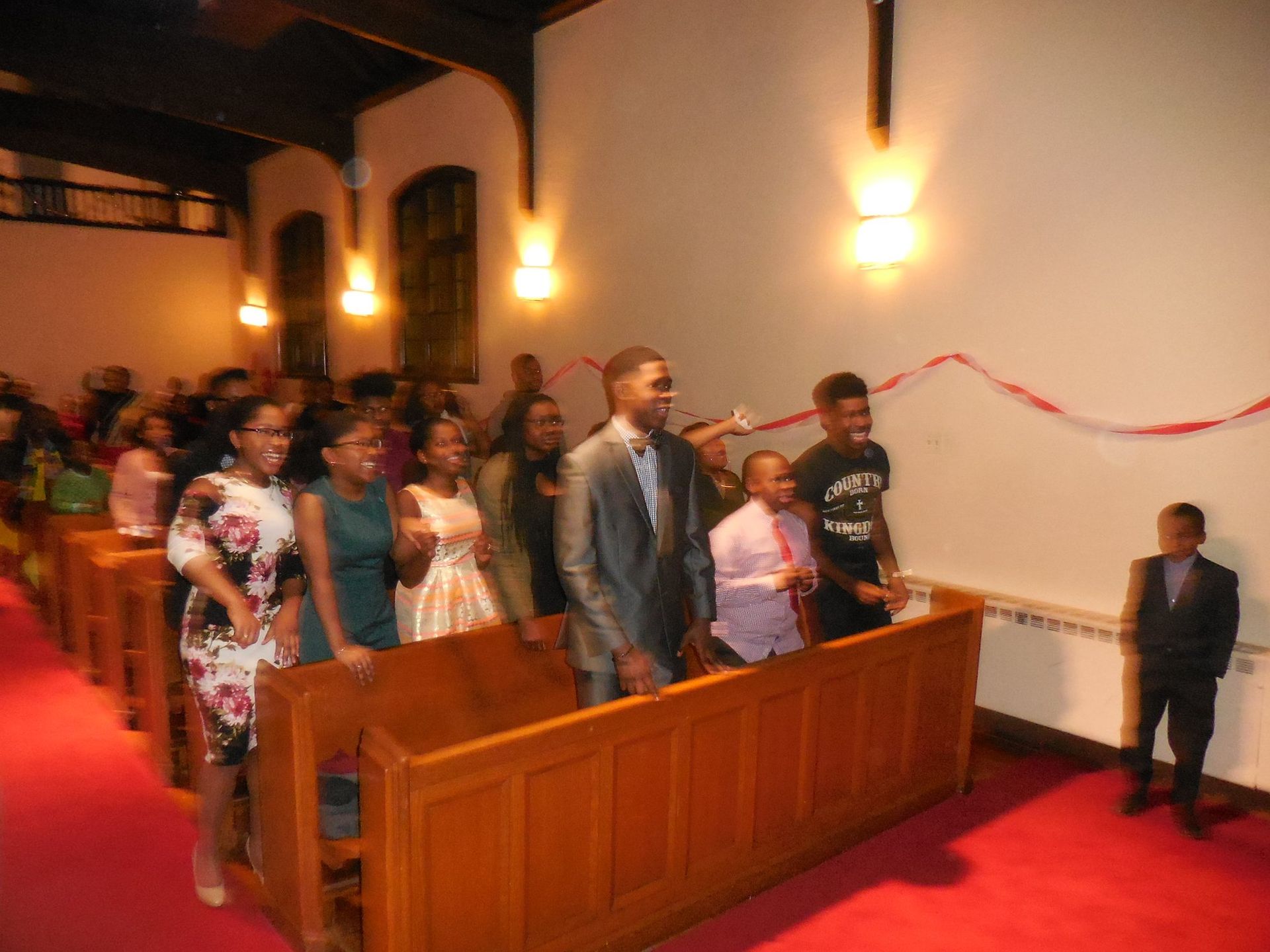 A diverse group in formal attire stands in rows along wooden pews inside a church, some smiling, facing forward.