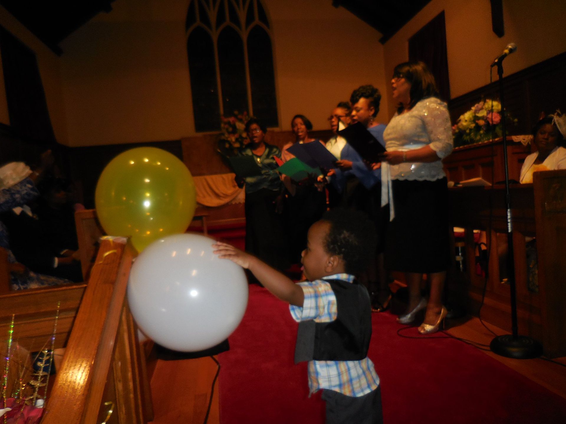 A child touches a white balloon in a church aisle as a group sings near the altar.