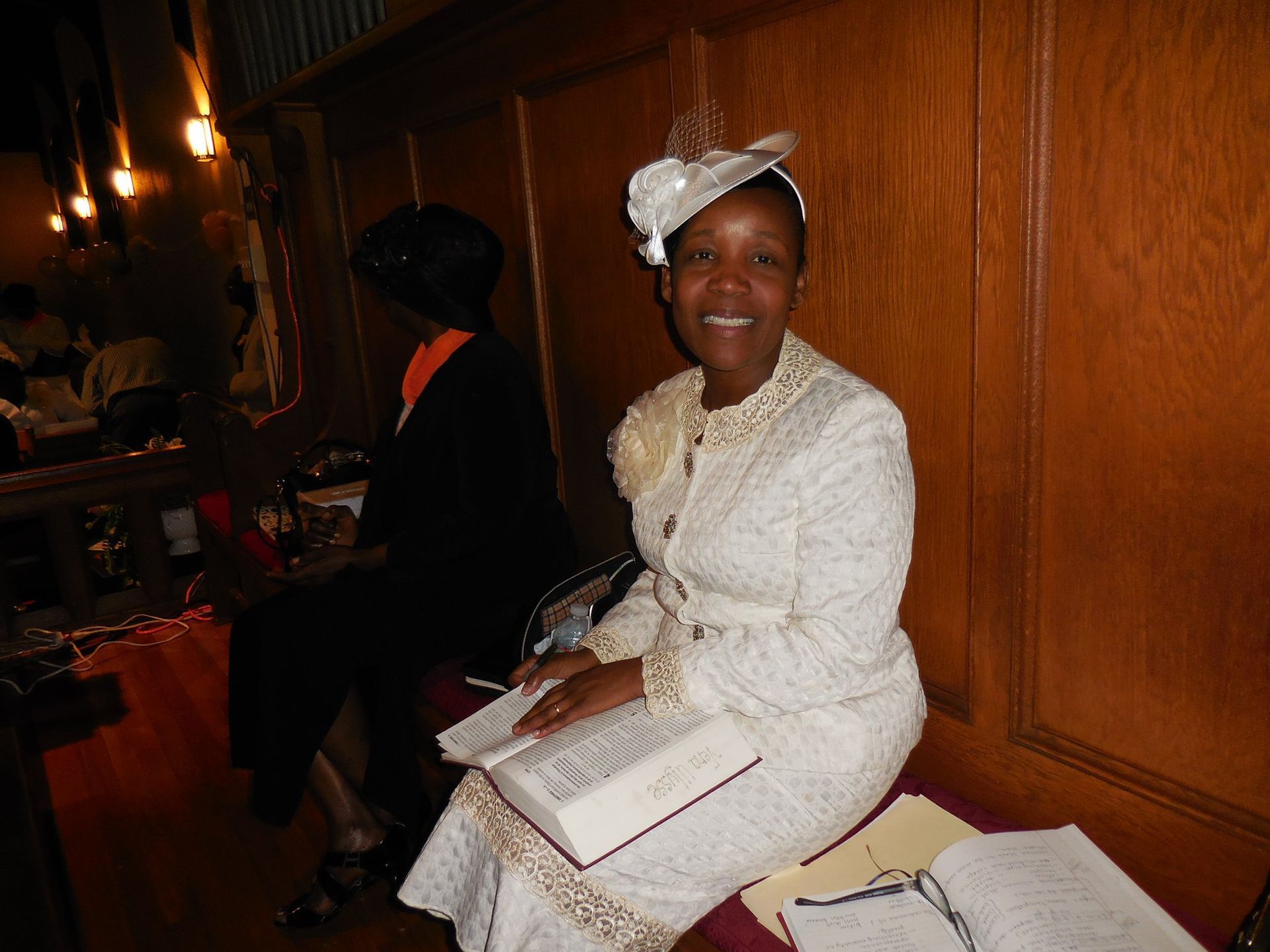 A smiling person wearing a cream-colored suit and matching hat, sitting in a wooden pew with an open book on their lap.