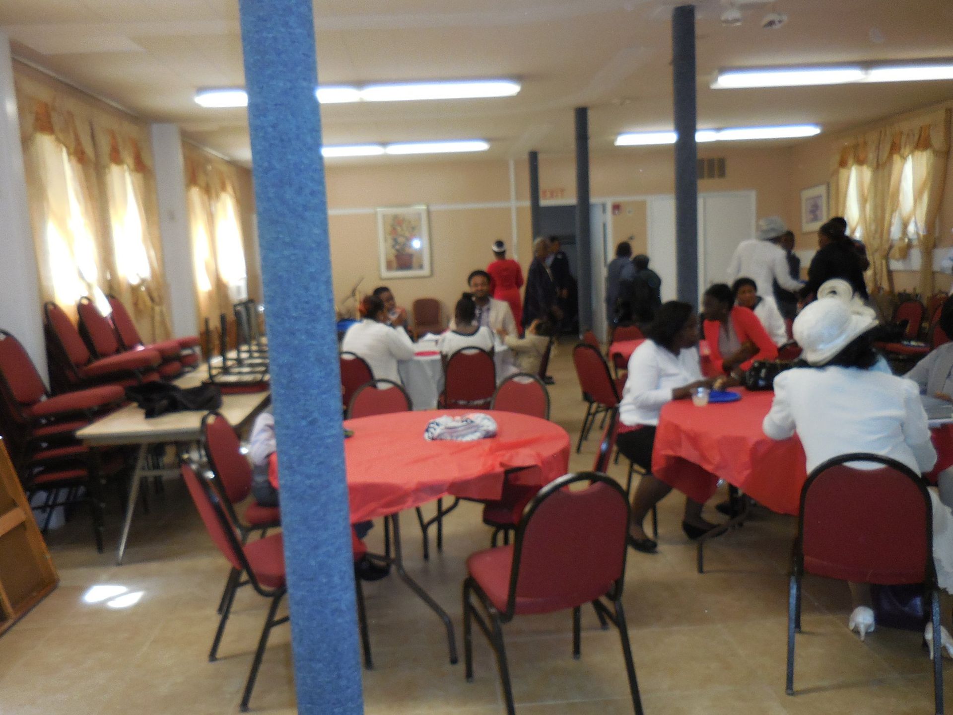 People gathered in a bright hall with red-clothed round tables, a blue pillar, and chairs in a social setting.